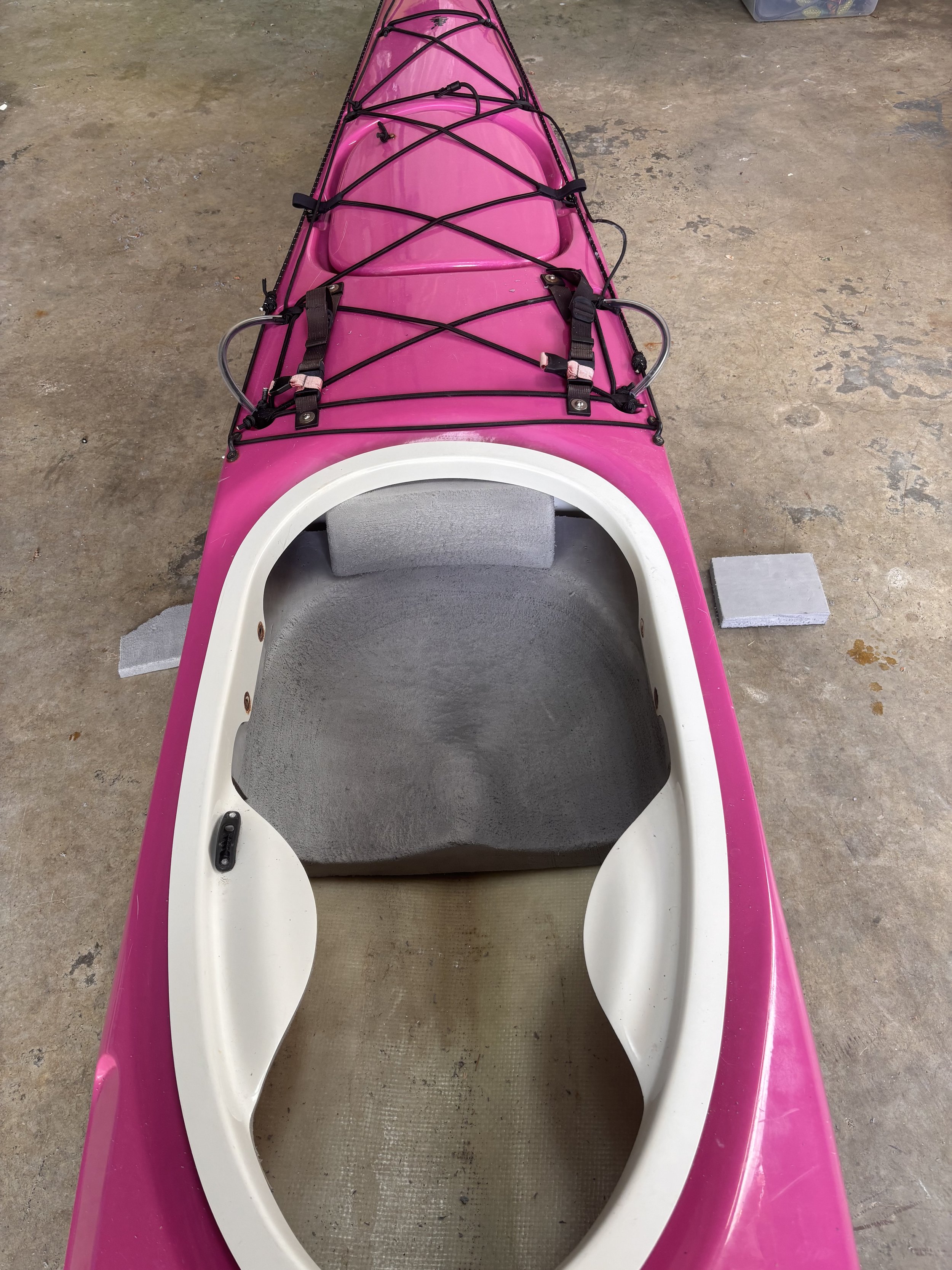 A pink kayak with a white circular opening in the front, situated on a concrete floor, with black cords and straps attached to the top.