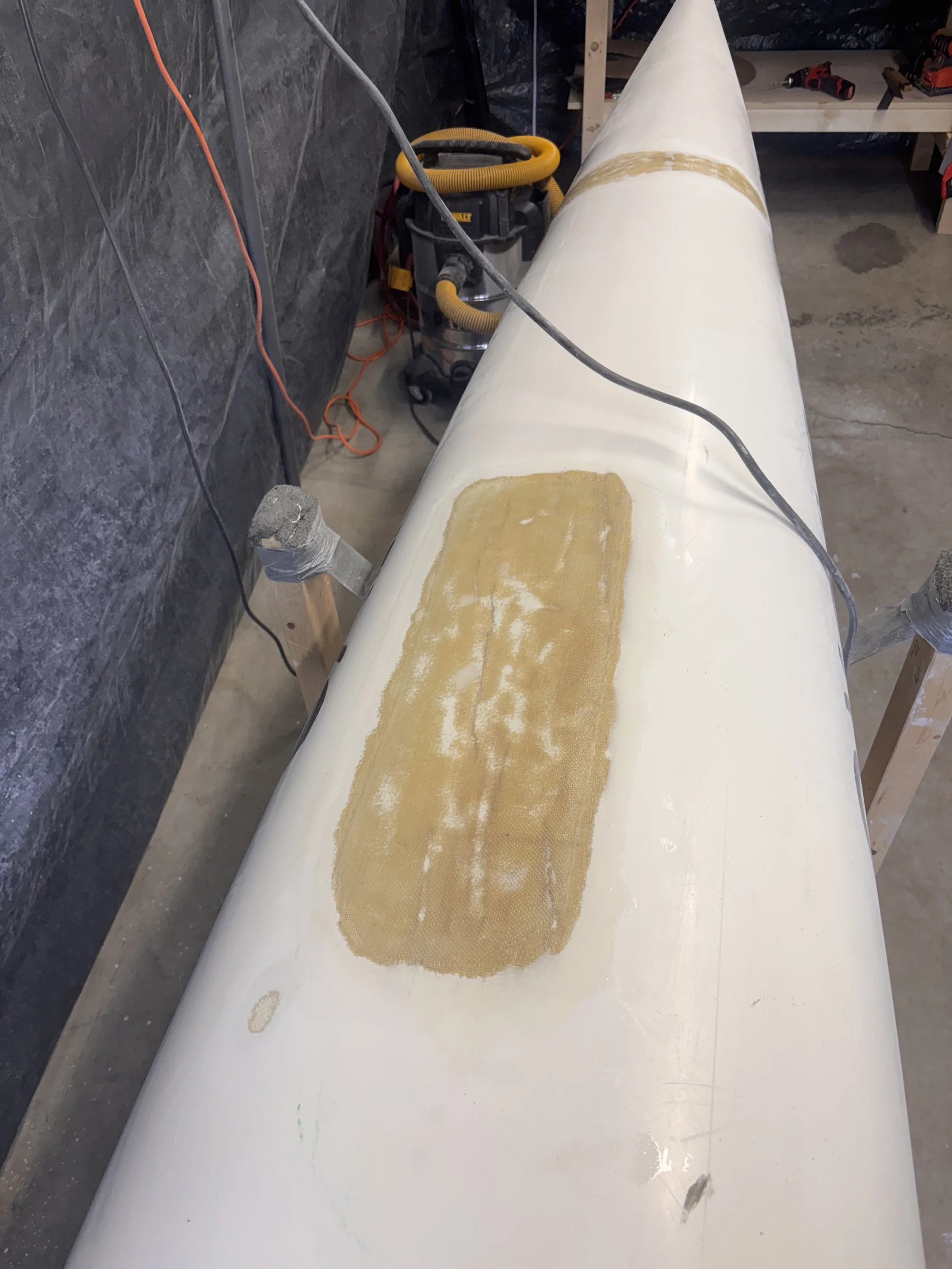 A large white cylindrical pipe in a workshop, with patches of foam or adhesive on its surface, surrounded by tools and equipment, including a vacuum cleaner and power drill.