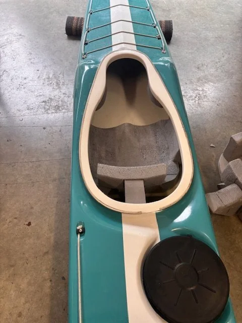 A kayak with a teal and white exterior, a black hatch cover at the front, and a seat inside, placed on a concrete floor.