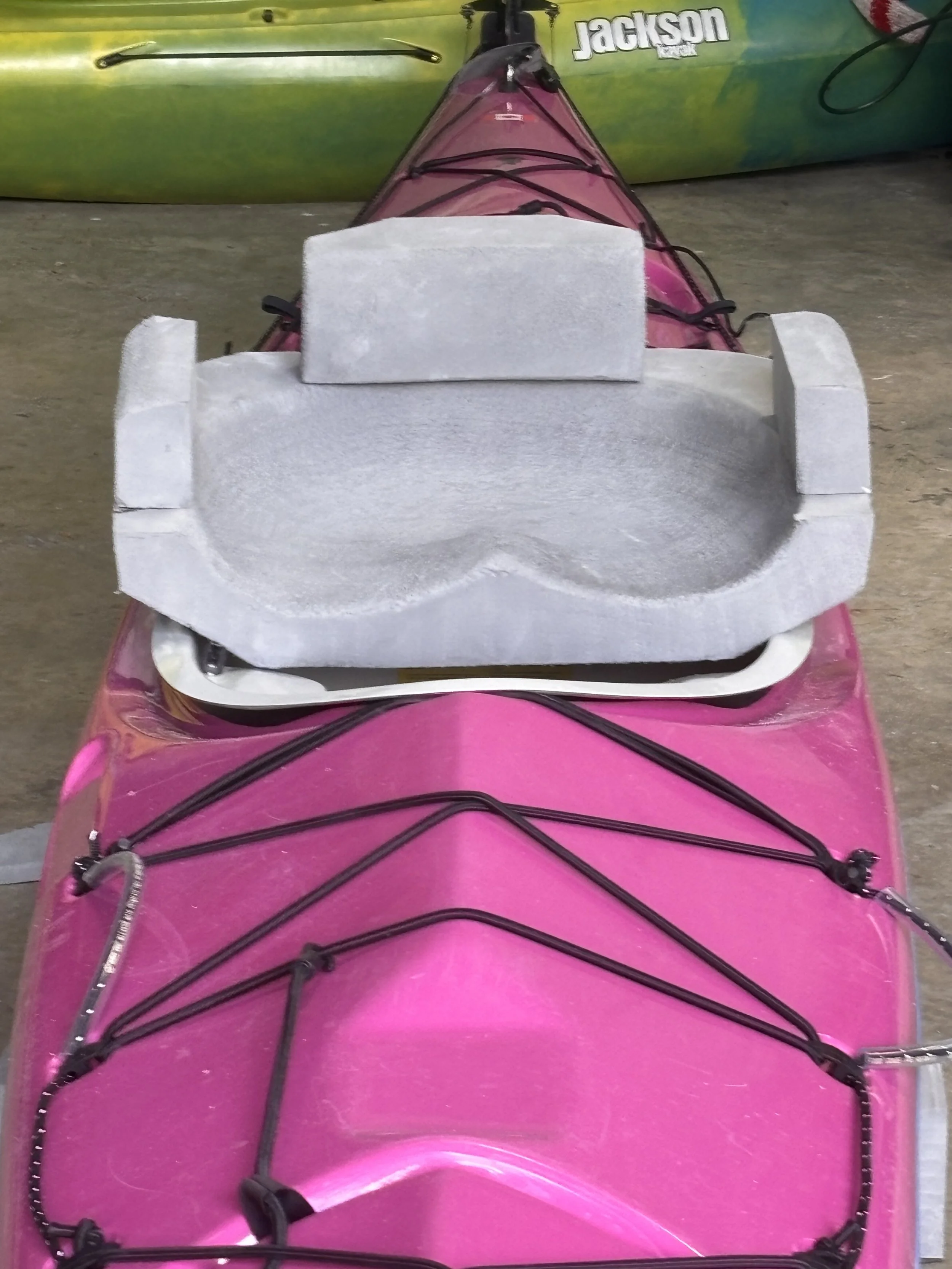 A pink kayak with black bungee cords tied on the front, placed on a concrete floor with another green kayak in the background.