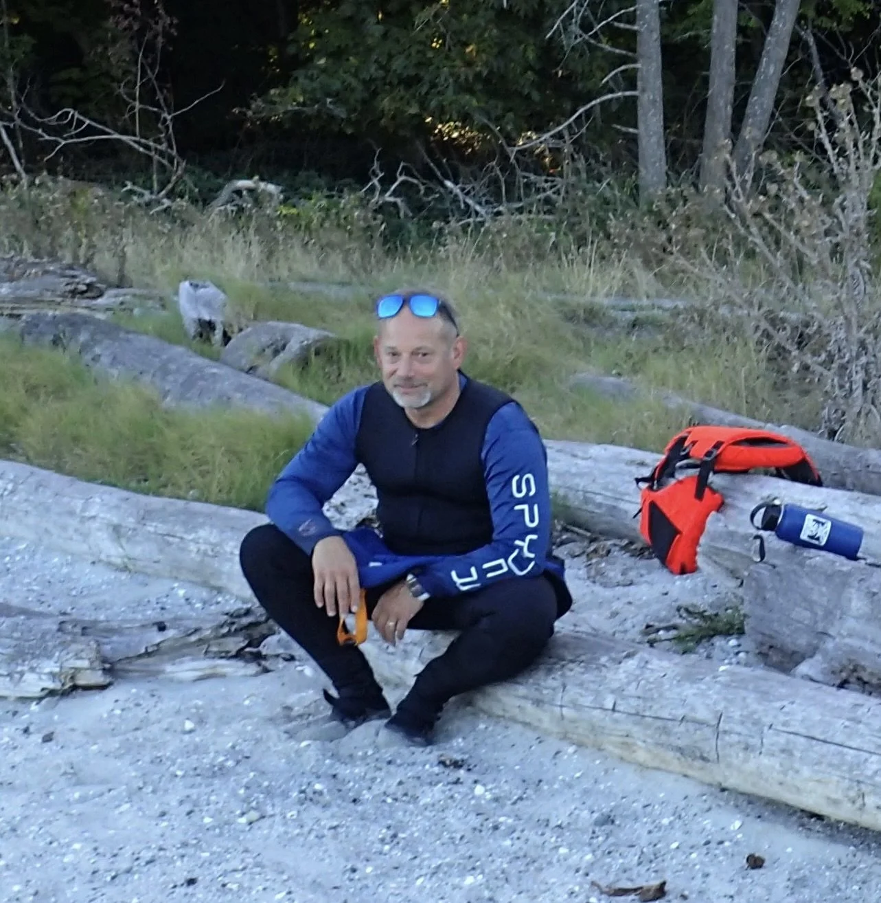 A man with short, light hair and a beard, dressed in a black and blue athletic outfit, squatting on a log at a trailside. There is a red backpack and a blue water bottle nearby on the log, with a green forest in the background.