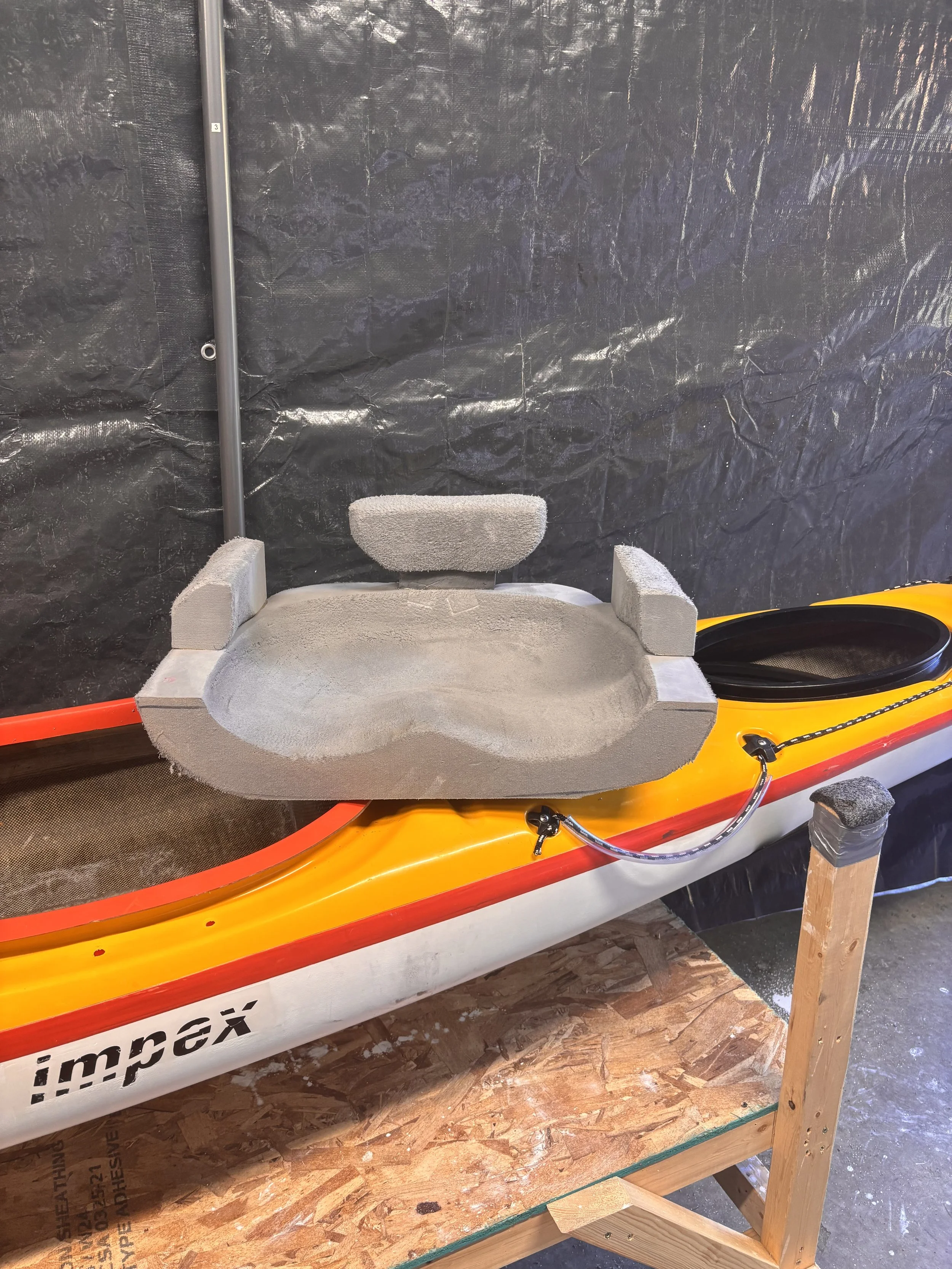 A small yellow kayak with a gray cushioned seat and a gray backrest, resting on a wooden stand in front of a black plastic tarp.