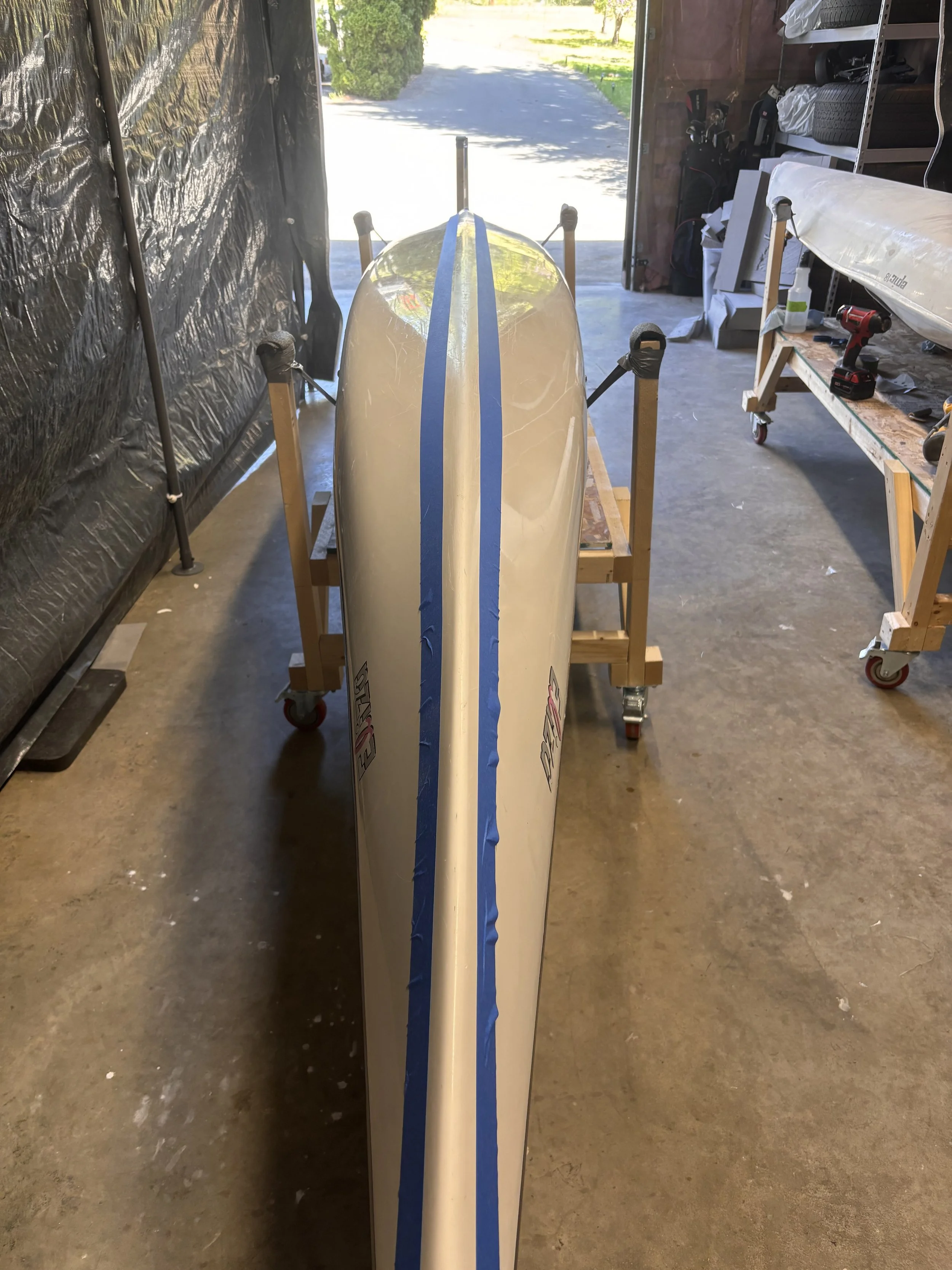 A boat inside a garage or workshop, with the front pointed toward an open garage door. The boat has blue painter's tape along the centerline. There are shelves with tires and tools to the right, and a workbench with a power drill and other equipment. The garage floor is concrete, and sunlight is coming in through the open door.