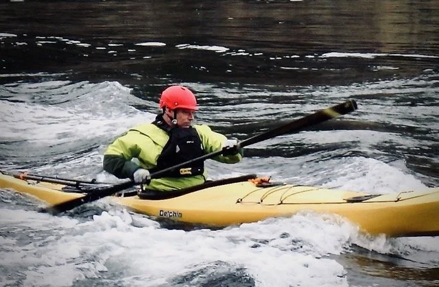 Person kayaking on a river, wearing a red helmet and yellow life jacket.