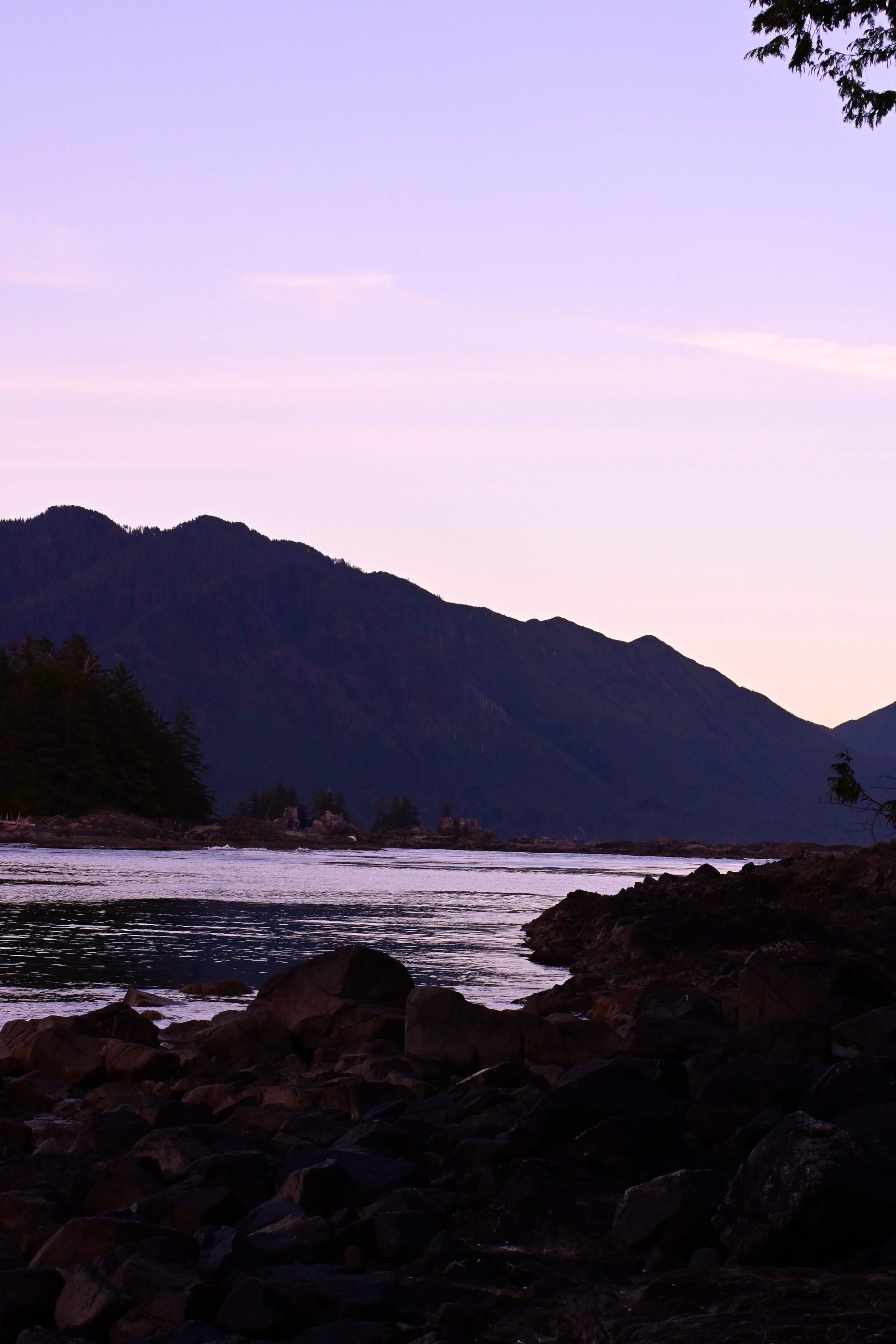 Scenic view of a river with rocks in the foreground, mountains in the background, and a pastel-colored sky at dusk or dawn.