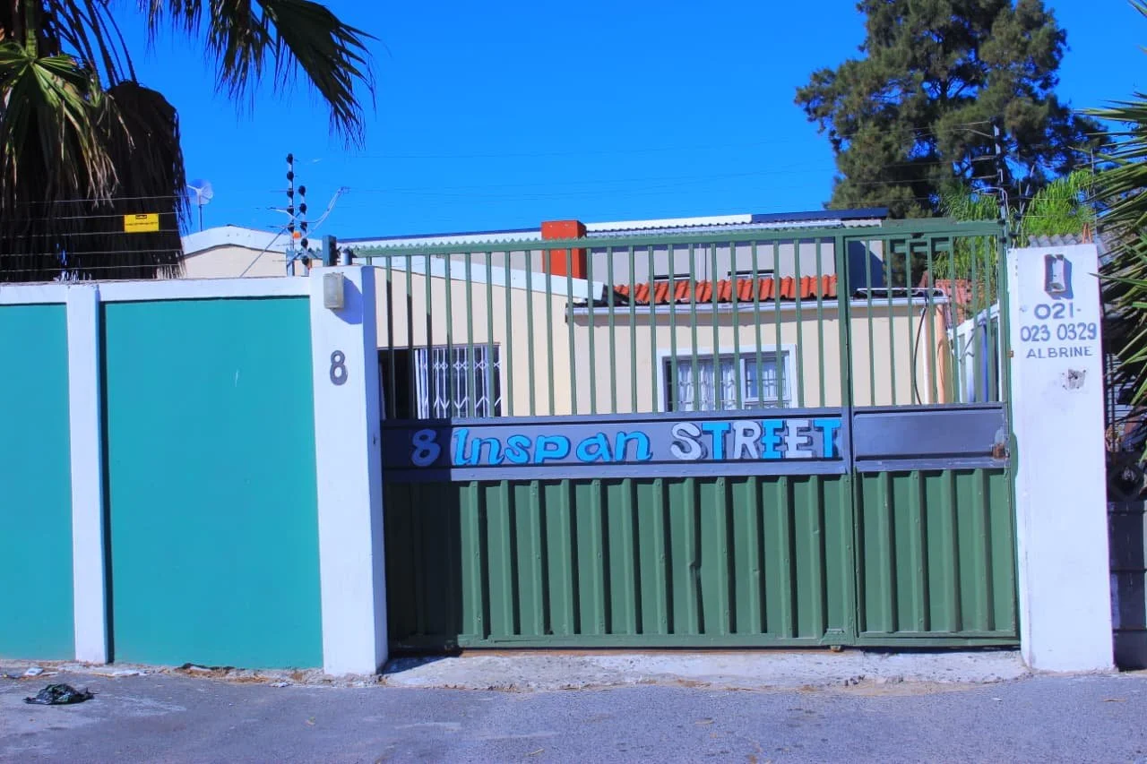 A green metal gate with a sign that reads "8 Inspan Street" in blue and white letters. The gate is set in a white wall, with a house and trees visible behind it. There are power lines above the gate and a blue sky.