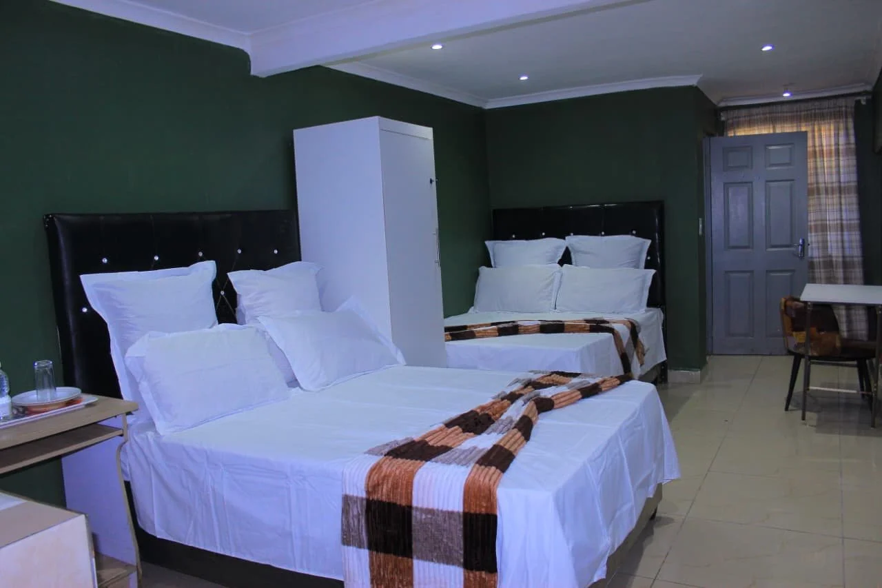 Hotel room with two beds, black headboards, white bedding, a wardrobe, a table with glasses and plates, and a door with a plaid curtain.