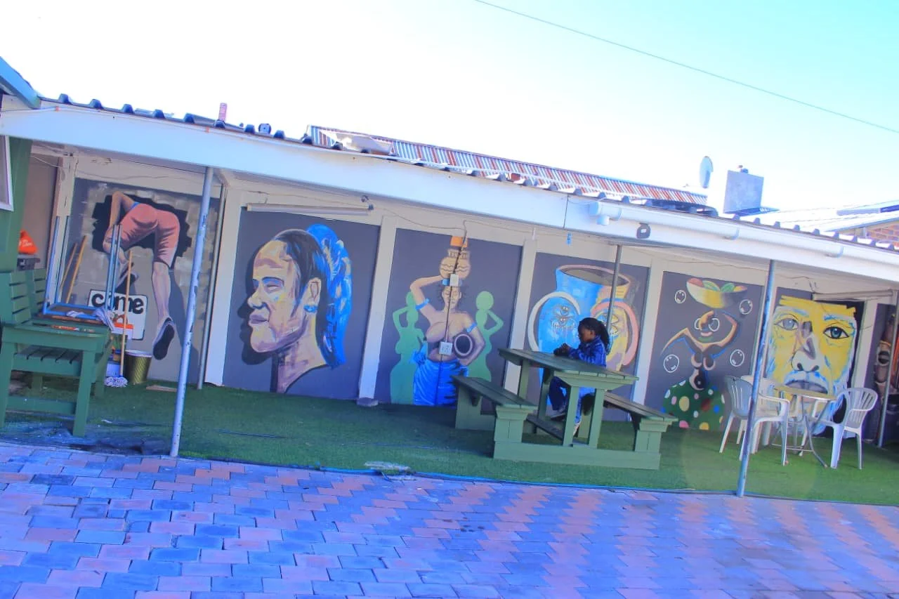 Colorful outdoor mural art on a building wall, featuring various portraits and abstract designs, with a person sitting at a picnic table and outdoor furniture.