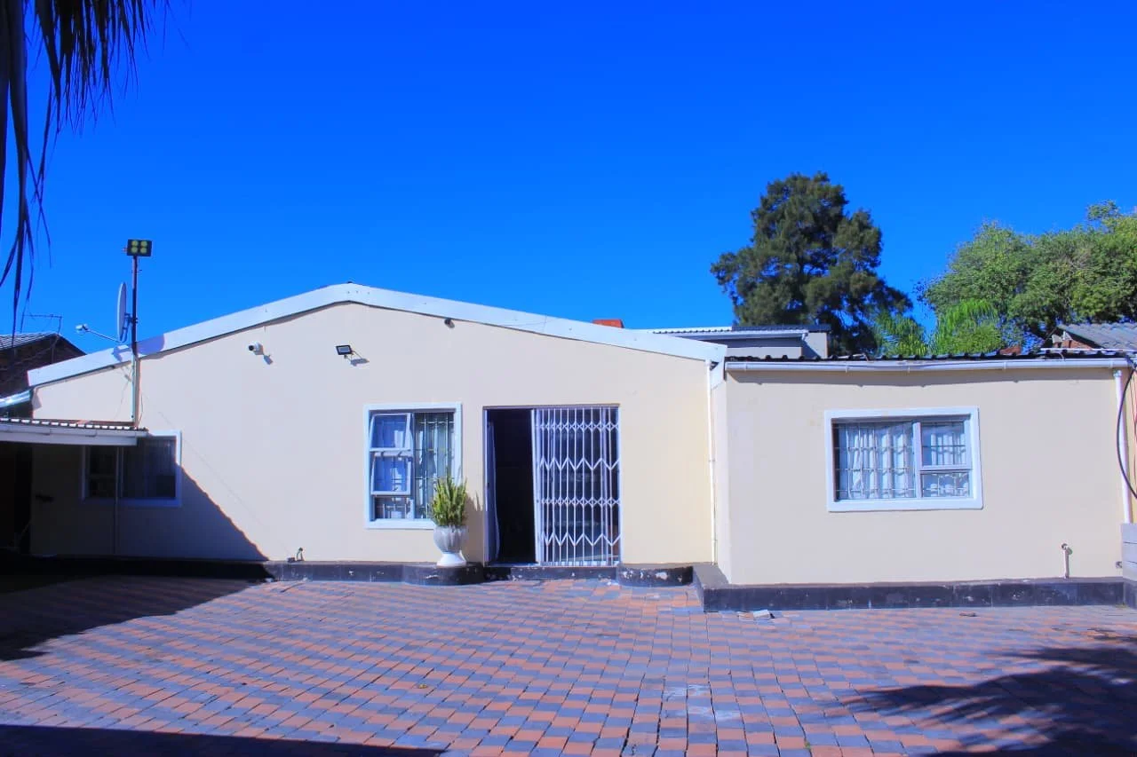 Single-story house with beige walls, white-framed windows with security bars, a metal gate, and a potted plant near the entrance. The house has a paved courtyard and a clear blue sky.