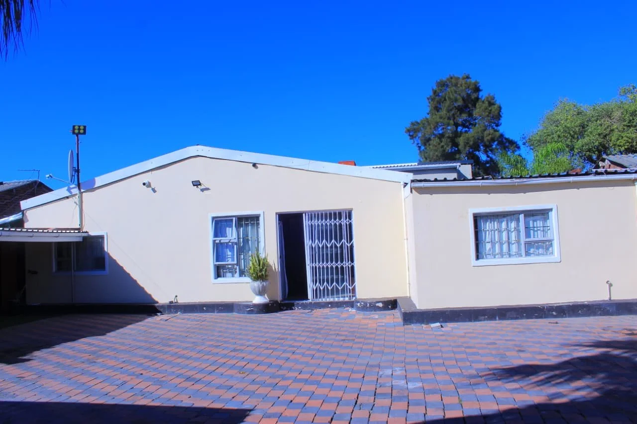 Front view of a single-story house with light yellow walls, white window frames, and a metal gate, surrounded by a brick driveway and green trees under a clear blue sky.