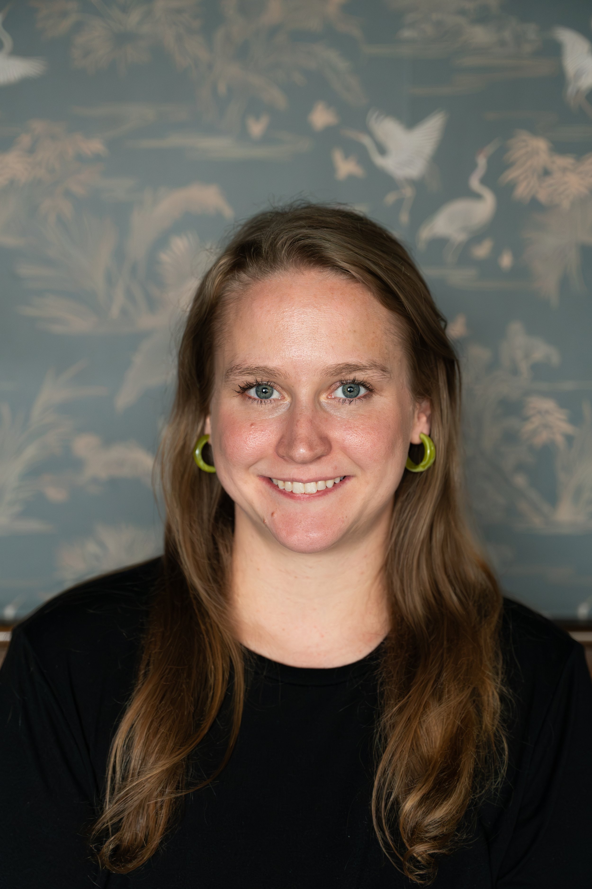 Portrait of a woman with long wavy brown hair, wearing lime green earrings and a black top, smiling in front of a blue wall with a white bird and foliage pattern.