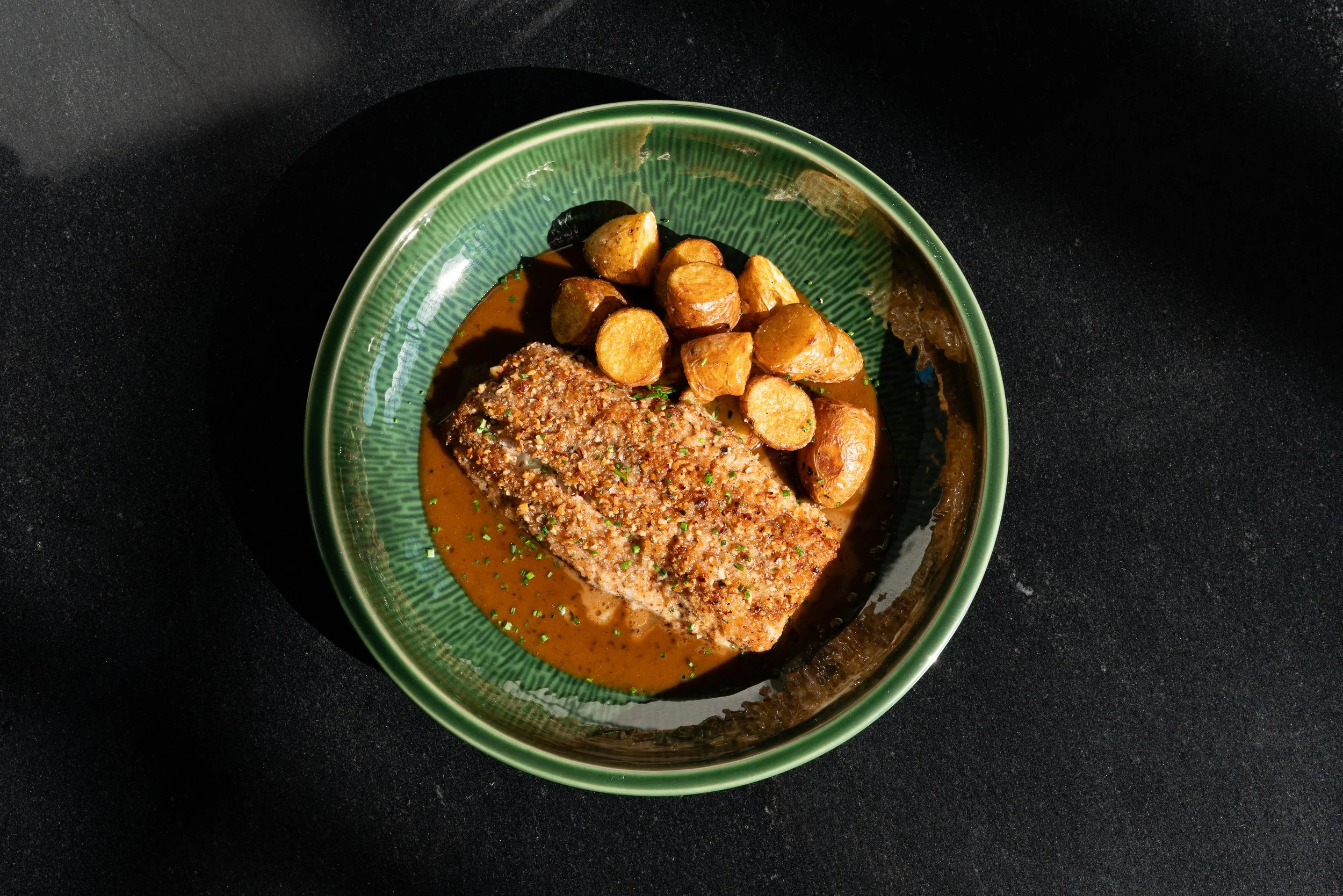 A green bowl contains a cooked fish fillet topped with seasoning, served with roasted potatoes in a sauce, garnished with chopped herbs, on a dark countertop.