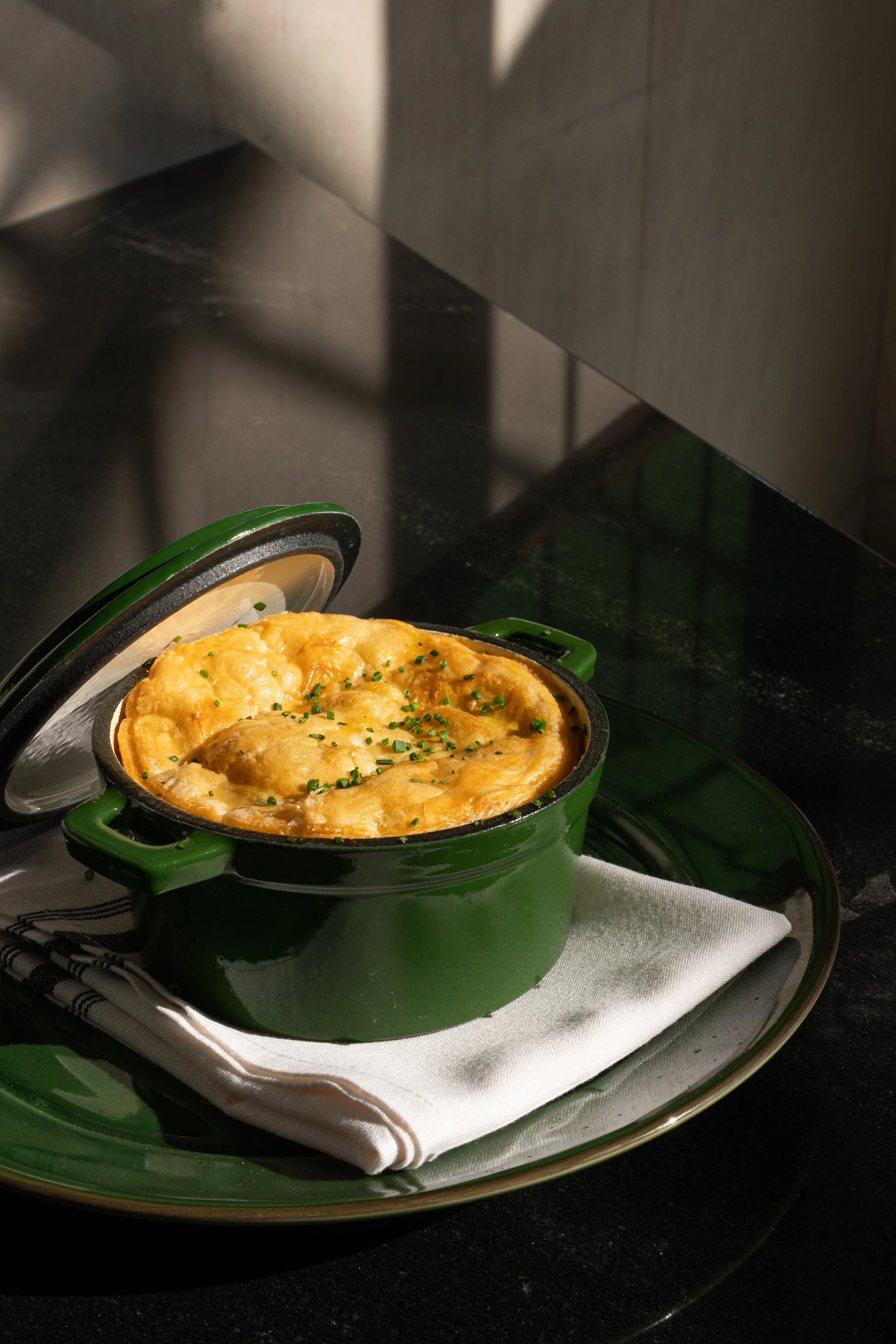 Baked casserole, in a green ceramic dish with a lid partially open, garnished with chopped herbs, on a green plate with a white napkin underneath.