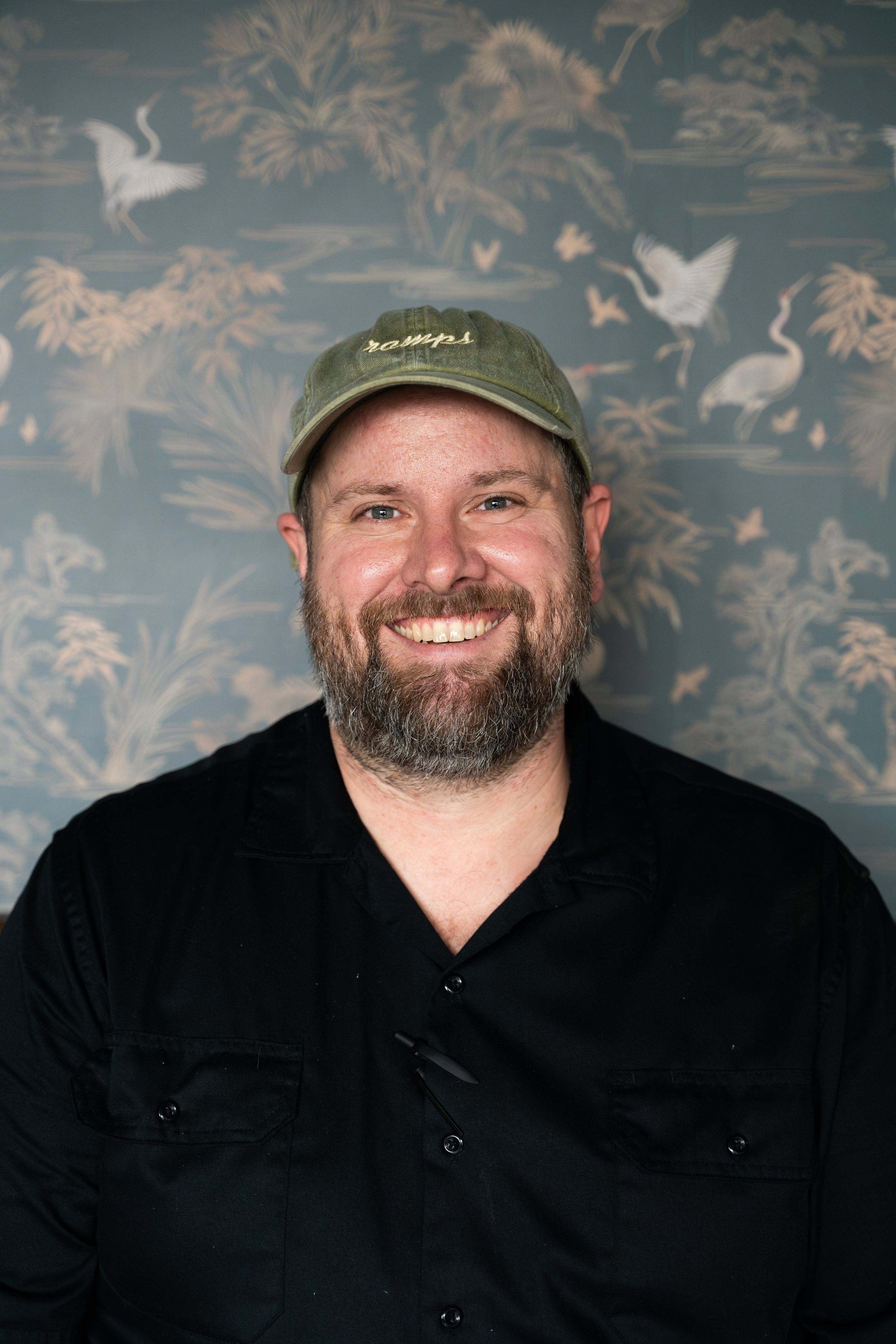 A smiling man with a beard and blue eyes, wearing a green baseball cap and a black shirt, standing in front of a blue wallpaper with white crane and nature patterns.