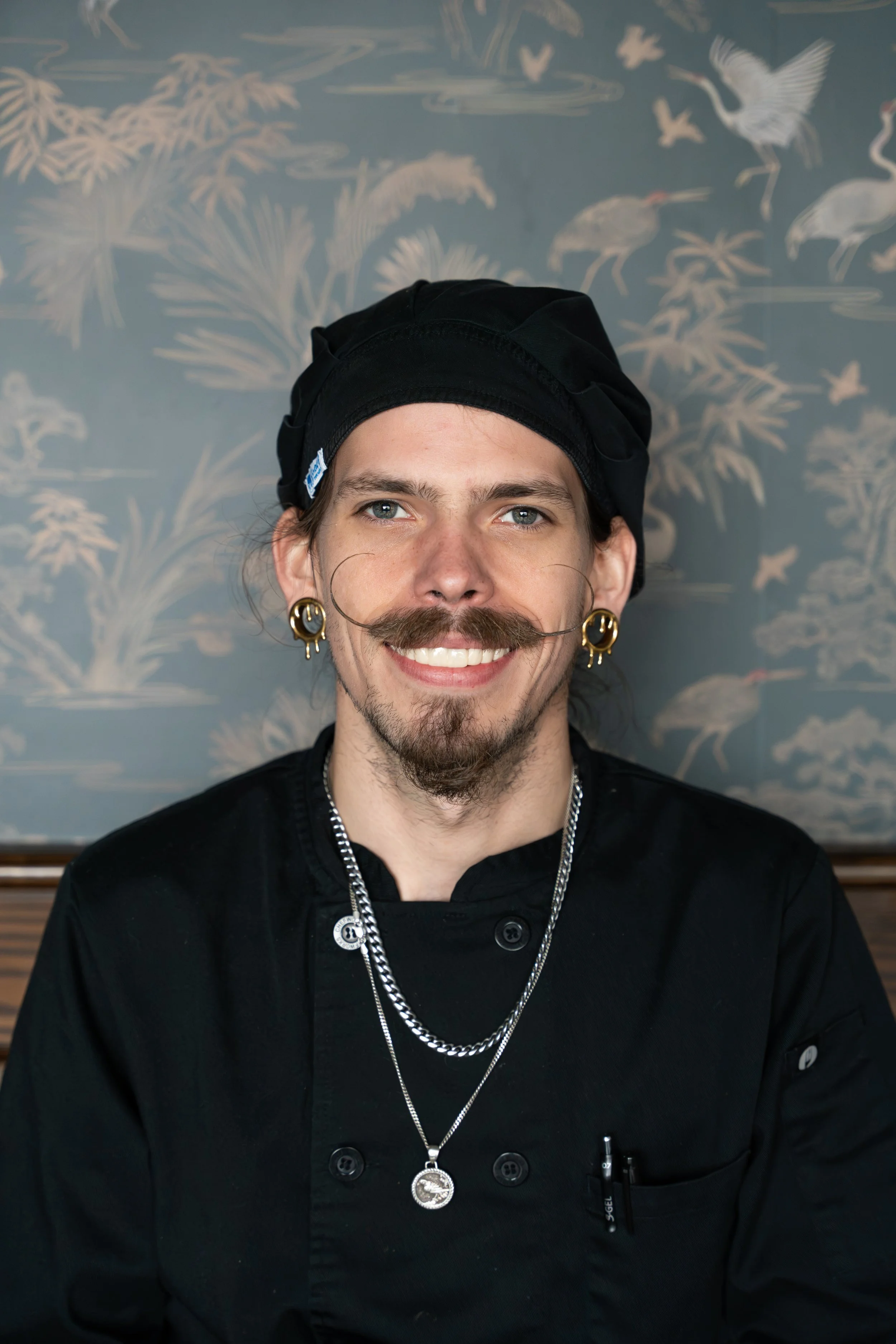 A smiling man with a mustache and goatee, wearing a black chef's coat, layered necklaces, gold earrings, and a black chef's hat. Background features a blue wall with white illustrations of cranes, horses, and plants.