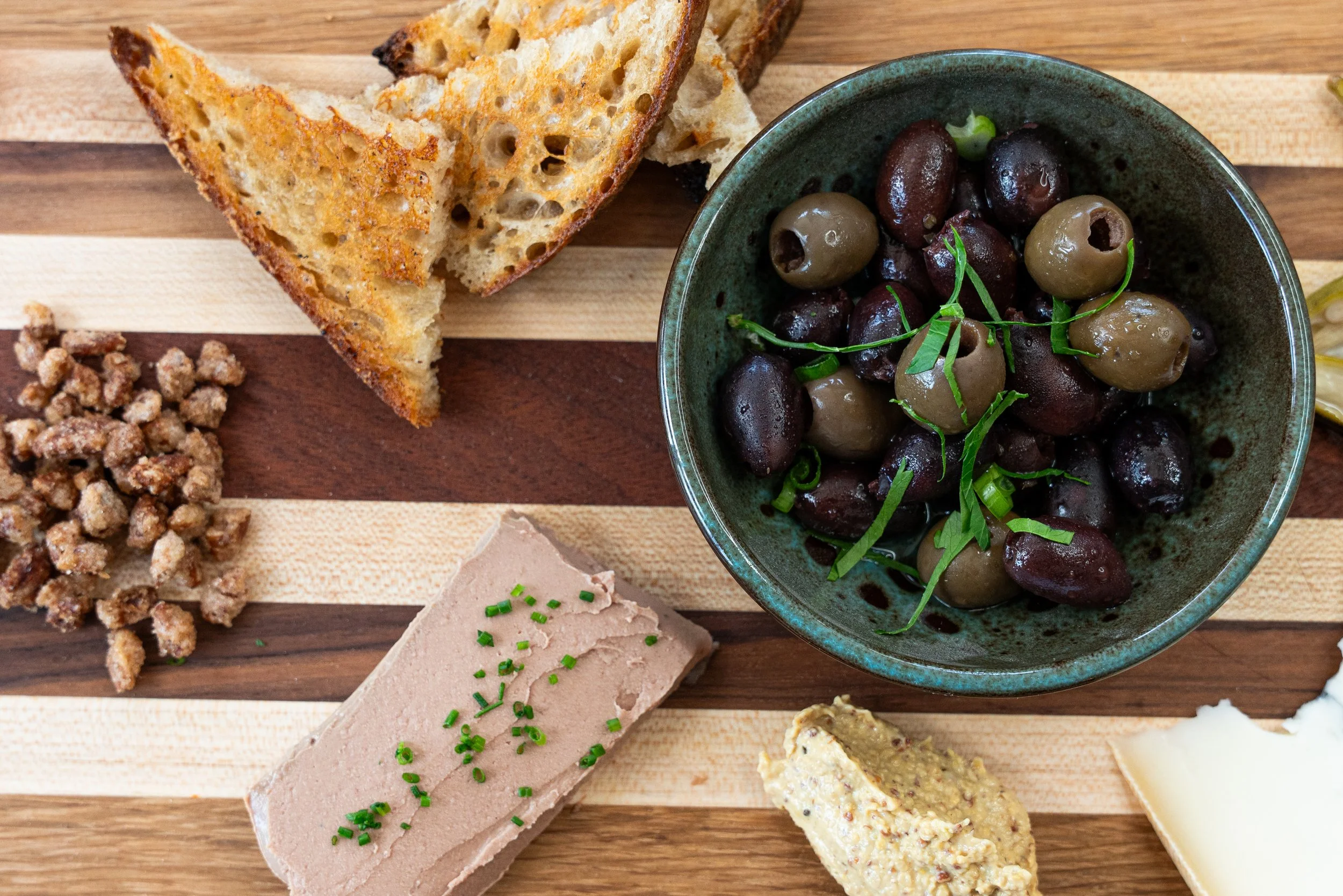 An assortment of appetizers on a wooden serving board including marinated olives in a green bowl, slices of pâté garnished with chives, slices of toasted bread, and various spreads and nuts.