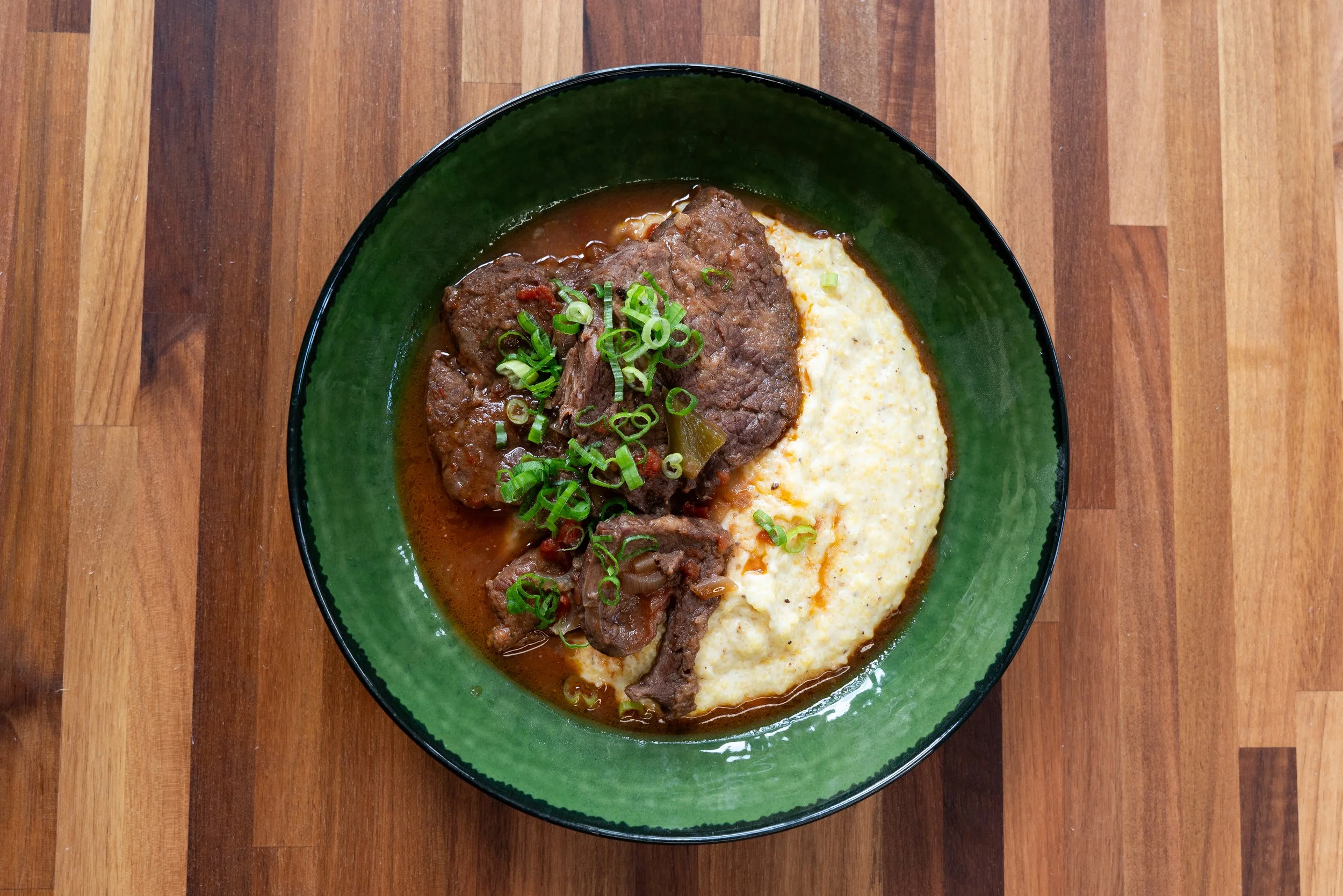 A bowl of beef stew with green onions served alongside creamy mashed potatoes, placed on a wooden table.