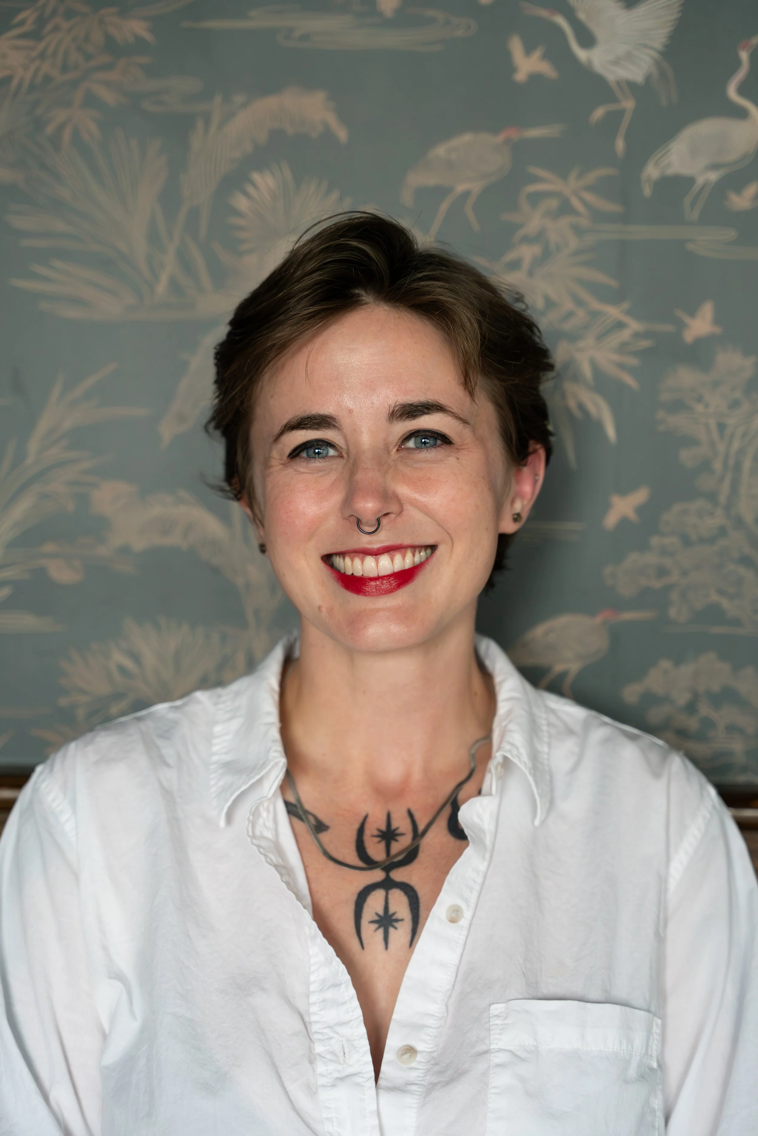 Close-up of a smiling woman with short brown hair, blue eyes, and a septum piercing, wearing a white shirt and a tattoo on her chest, standing in front of a patterned background with cranes and nature motifs.