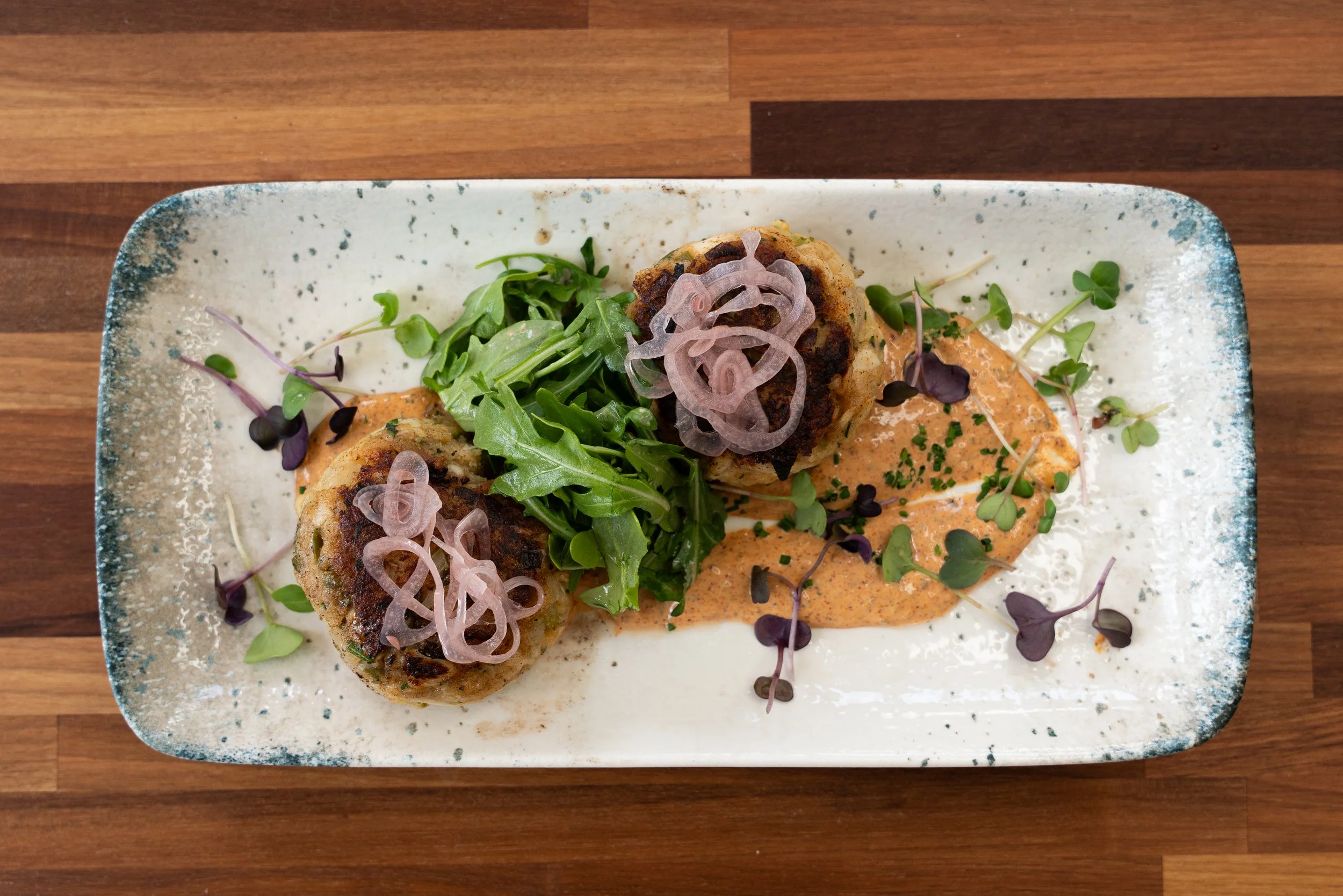 Plate of fried food garnished with greens, microgreens, and pickled onions, with a creamy sauce and spring herbs on a speckled ceramic plate on a wooden table.