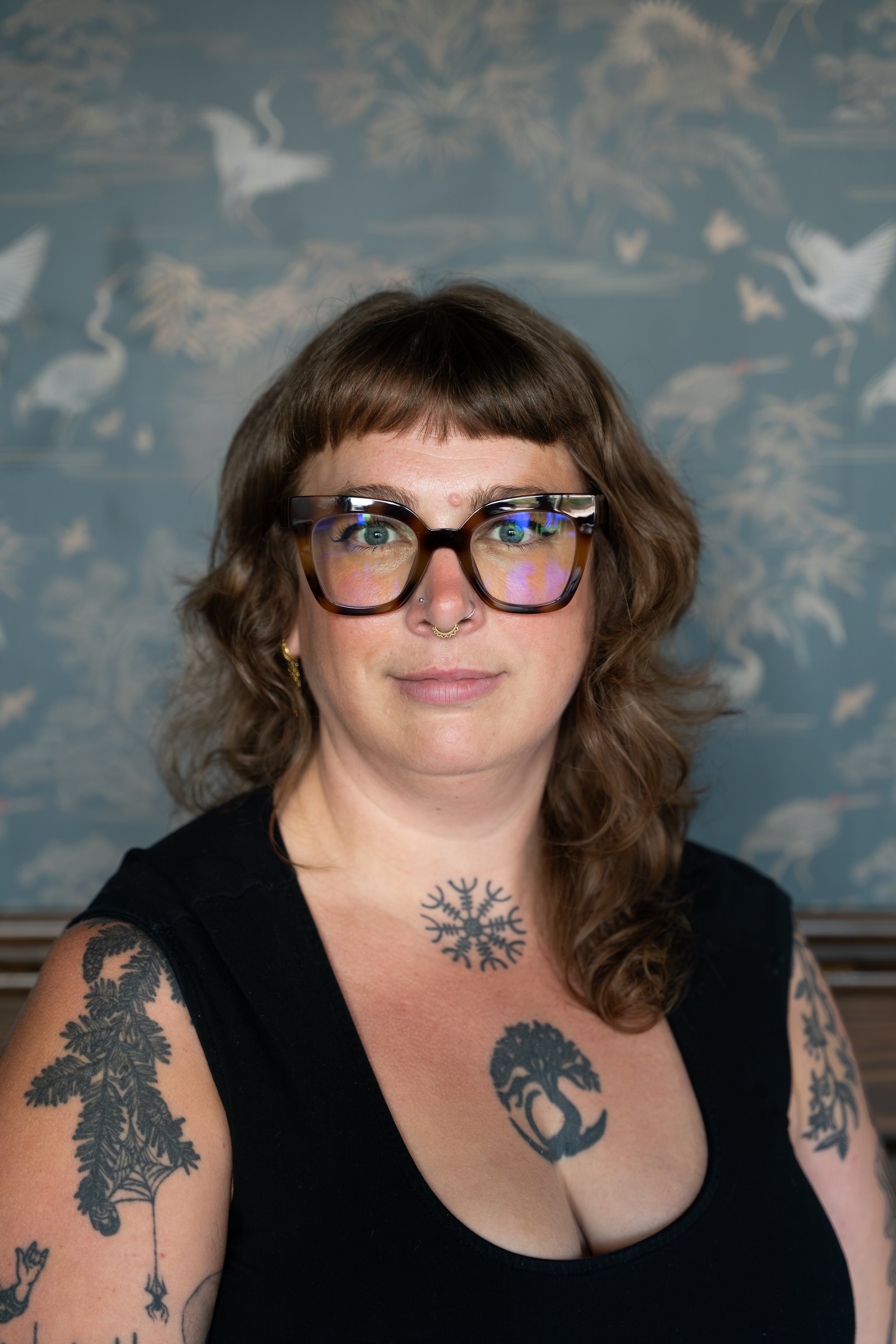 A woman with glasses, tattoos on her arms and chest, and a septum piercing, standing in front of a wallpaper with white birds and plants.