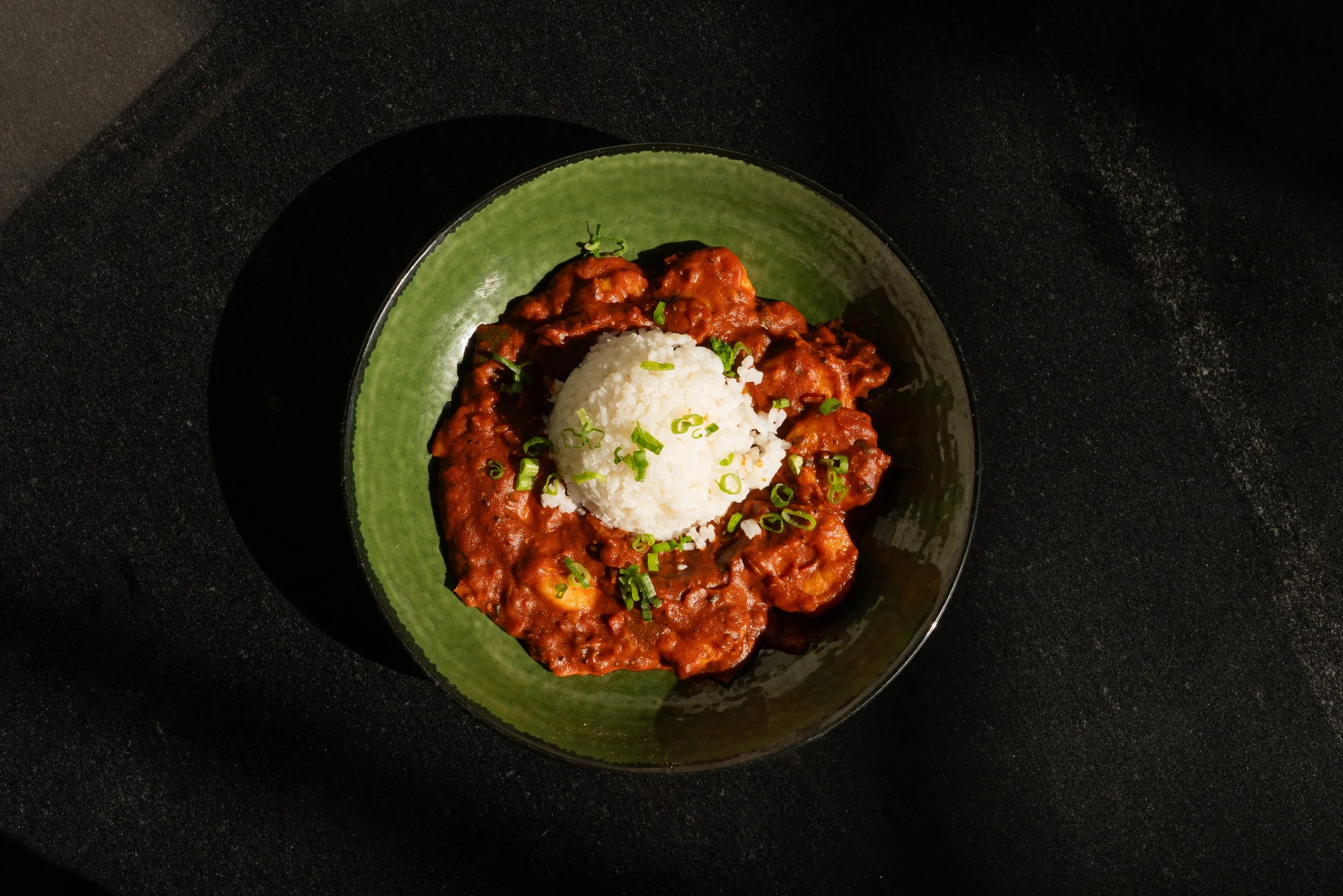 A black bowl with a green interior contains cooked meat in a reddish sauce, garnished with chopped green onions, and a portion of white rice on top.