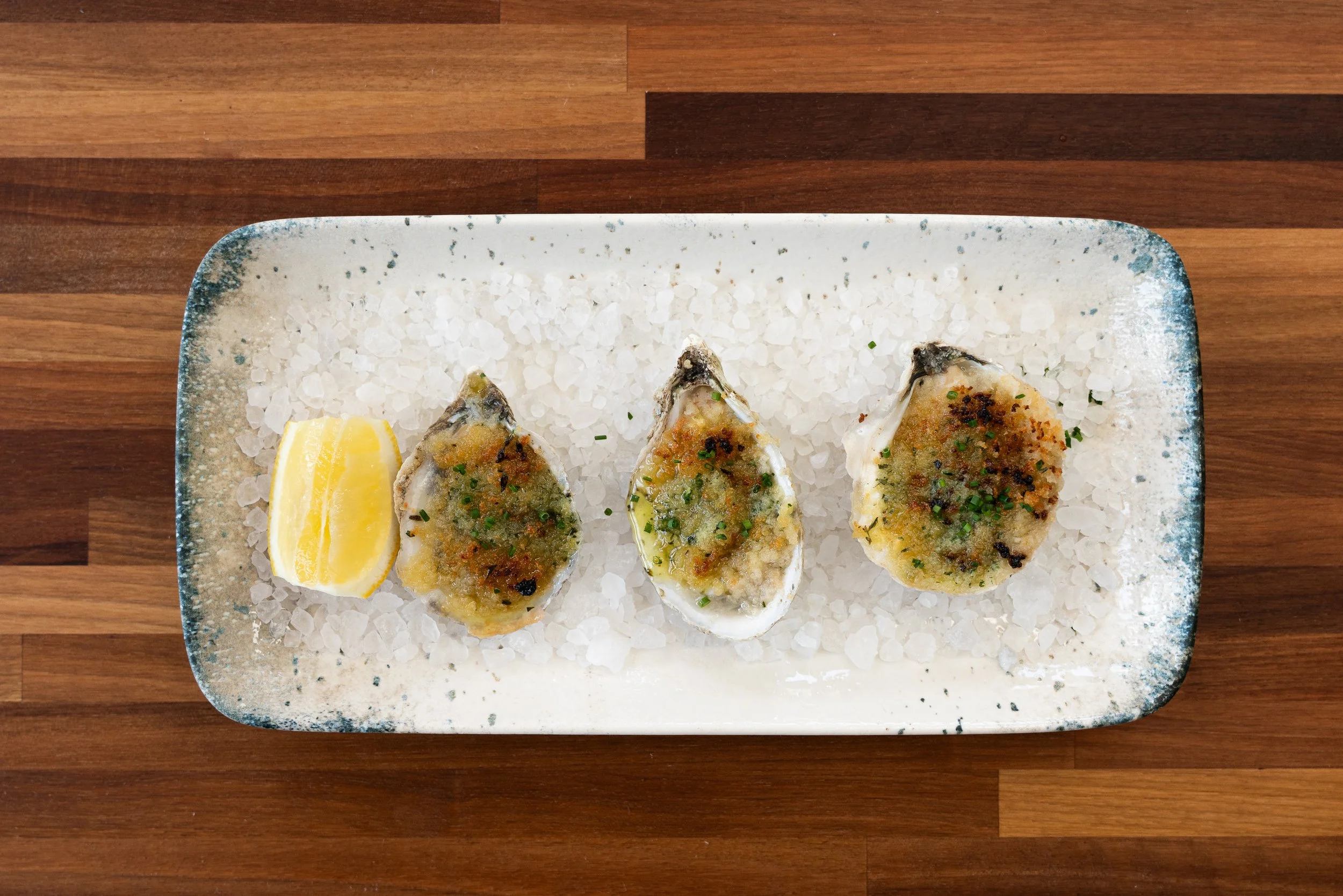 A rectangular plate with three baked oysters topped with herbs, a lemon wedge, and a bed of salt on a wooden table.