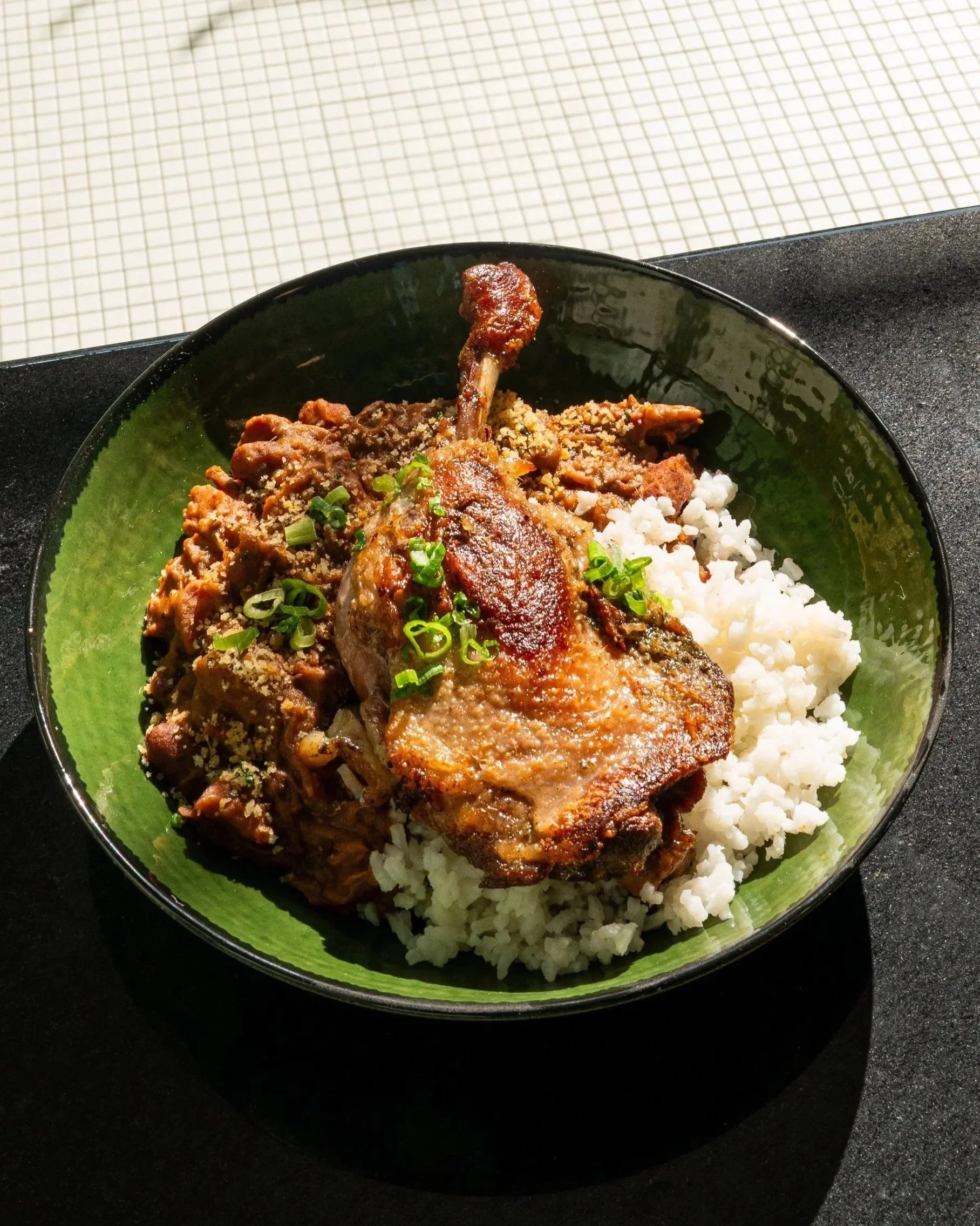 Close-up of a bowl of rice topped with cooked meat and garnished with chopped green onions, with a piece of chicken leg in the center, on a black surface.