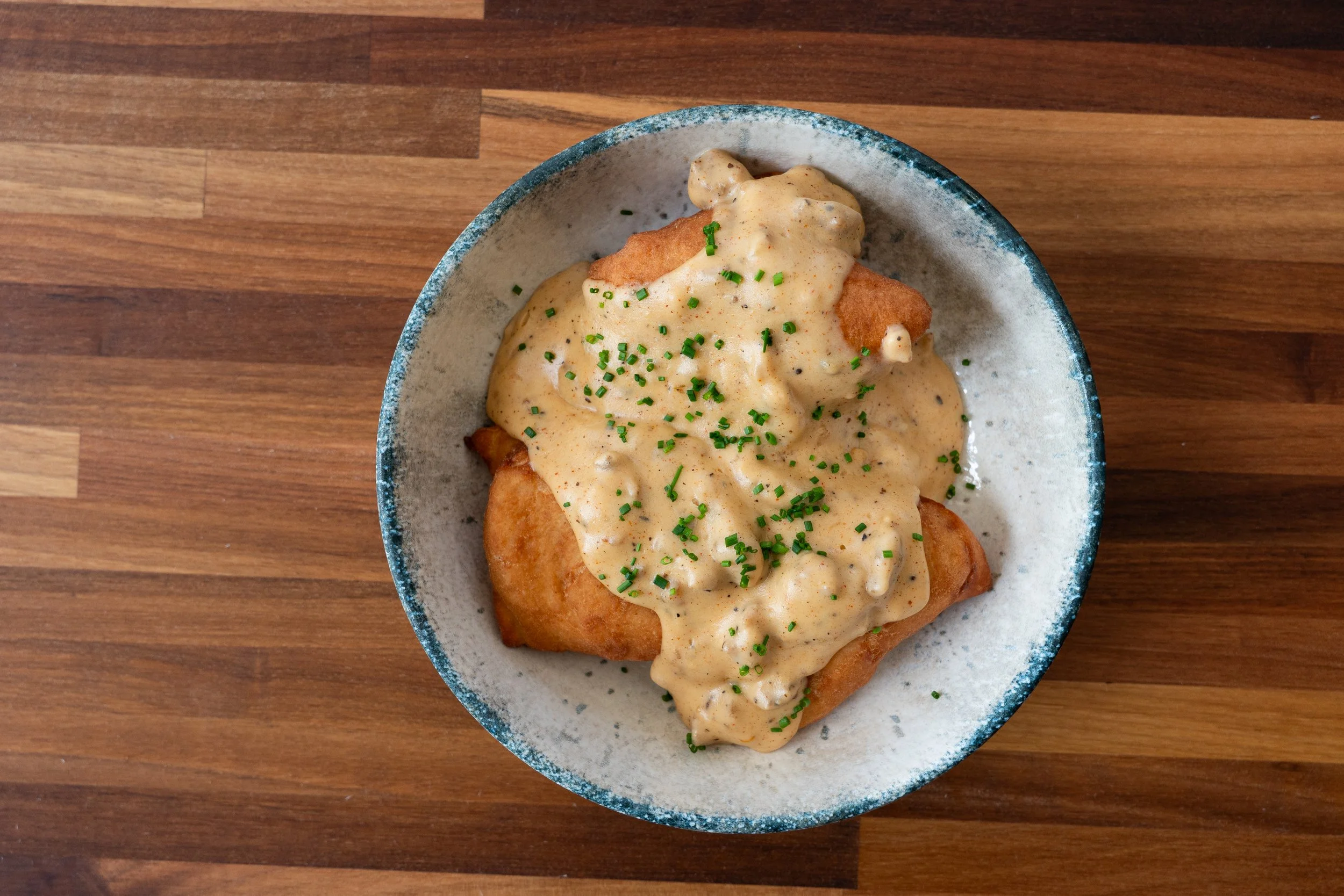 A bowl of fish and chips topped with creamy tartar sauce and chopped chives on a wooden table.