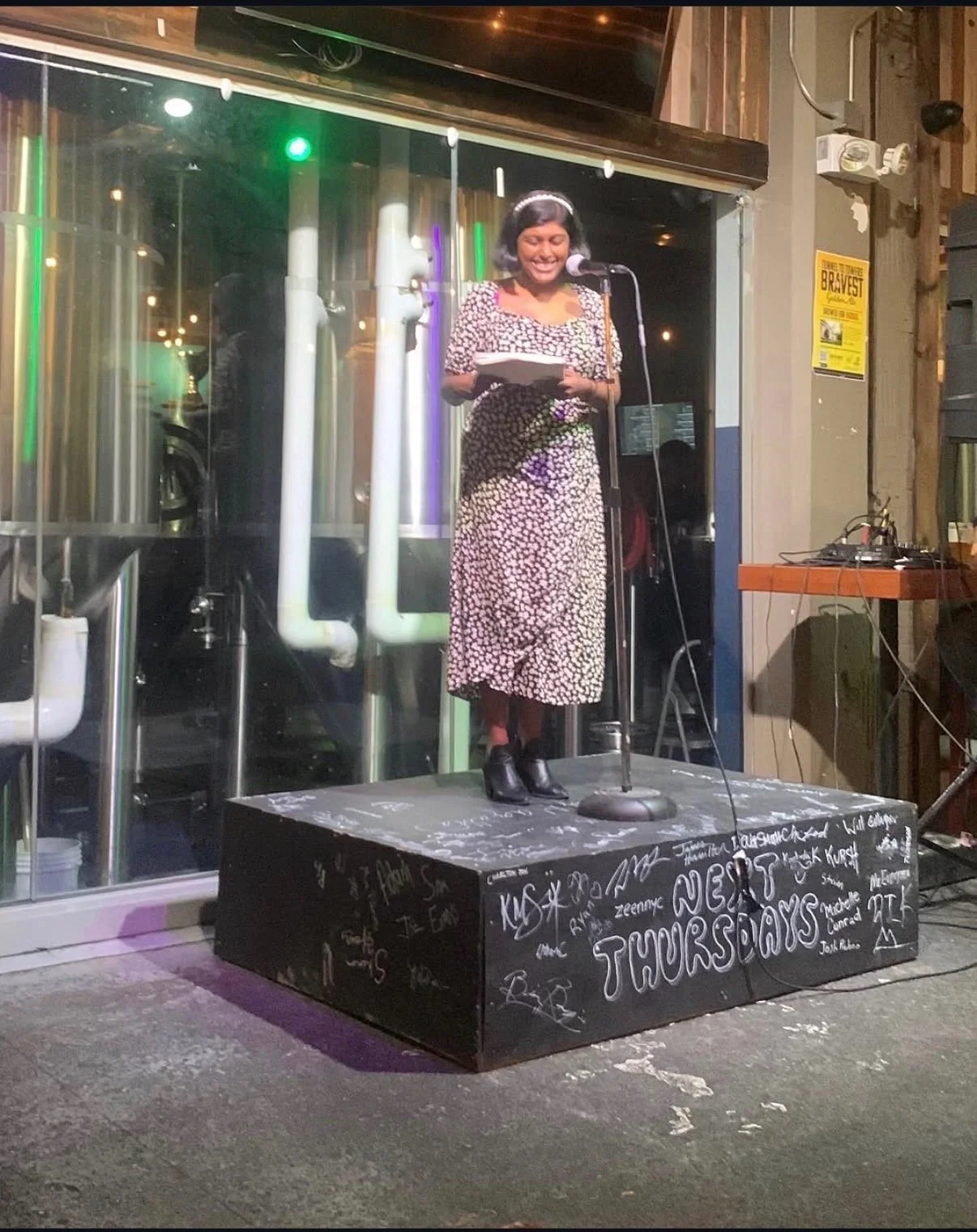 A woman with short dark hair, wearing a black and white floral dress, smiling while reading or speaking at a microphone on a small stage in a bar or restaurant. The stage has a black surface with white graffiti-style writing, including 'TONIGHT THURSDAYS.' Behind her, there are metallic pipes or industrial equipment, and to the right, a small table with audio equipment and wires.