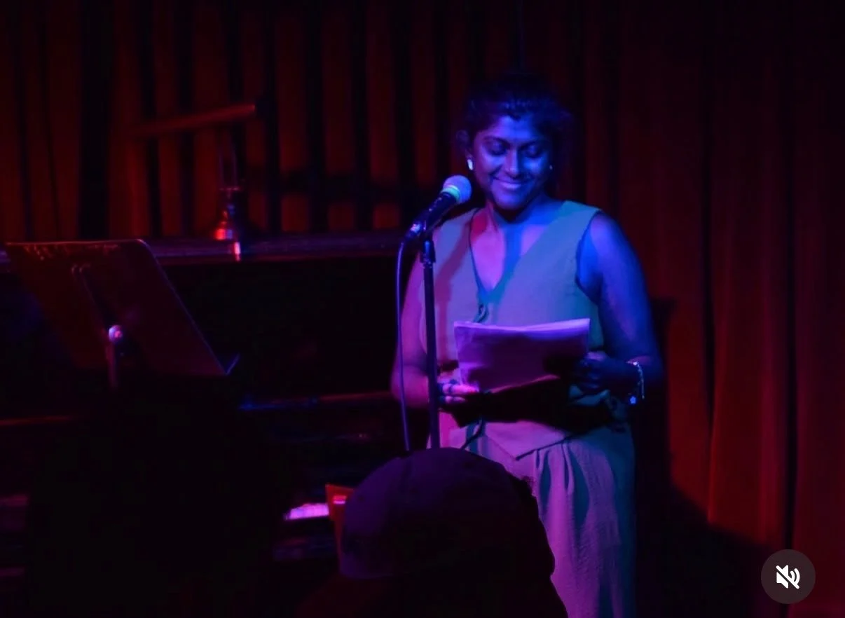 Woman speaking or performing with a piano in a dimly lit venue with red curtains.