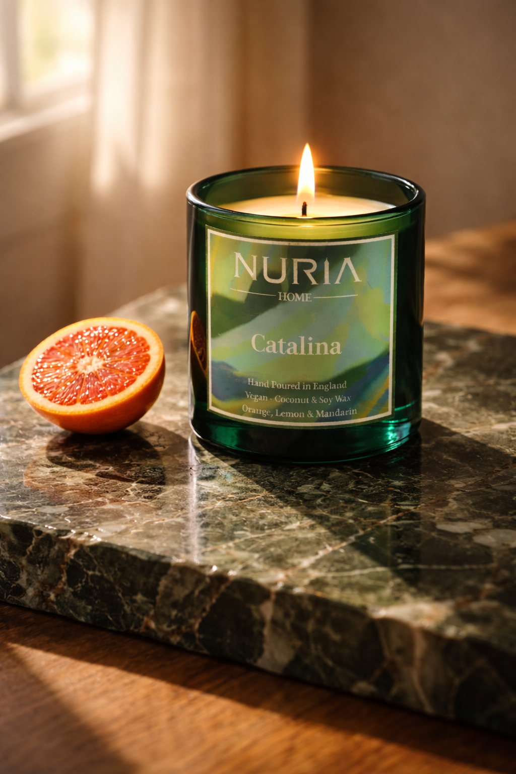 A lit candle in a green container labeled 'Nuria Home Catalina' placed on a marble surface next to half a grapefruit.