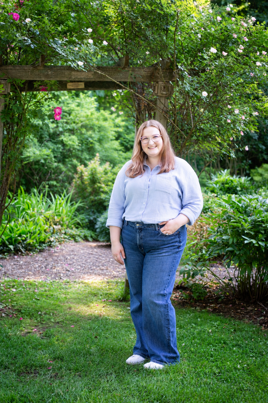 A woman smiling outdoors in a garden with lush green foliage, flowers, and a wooden archway.