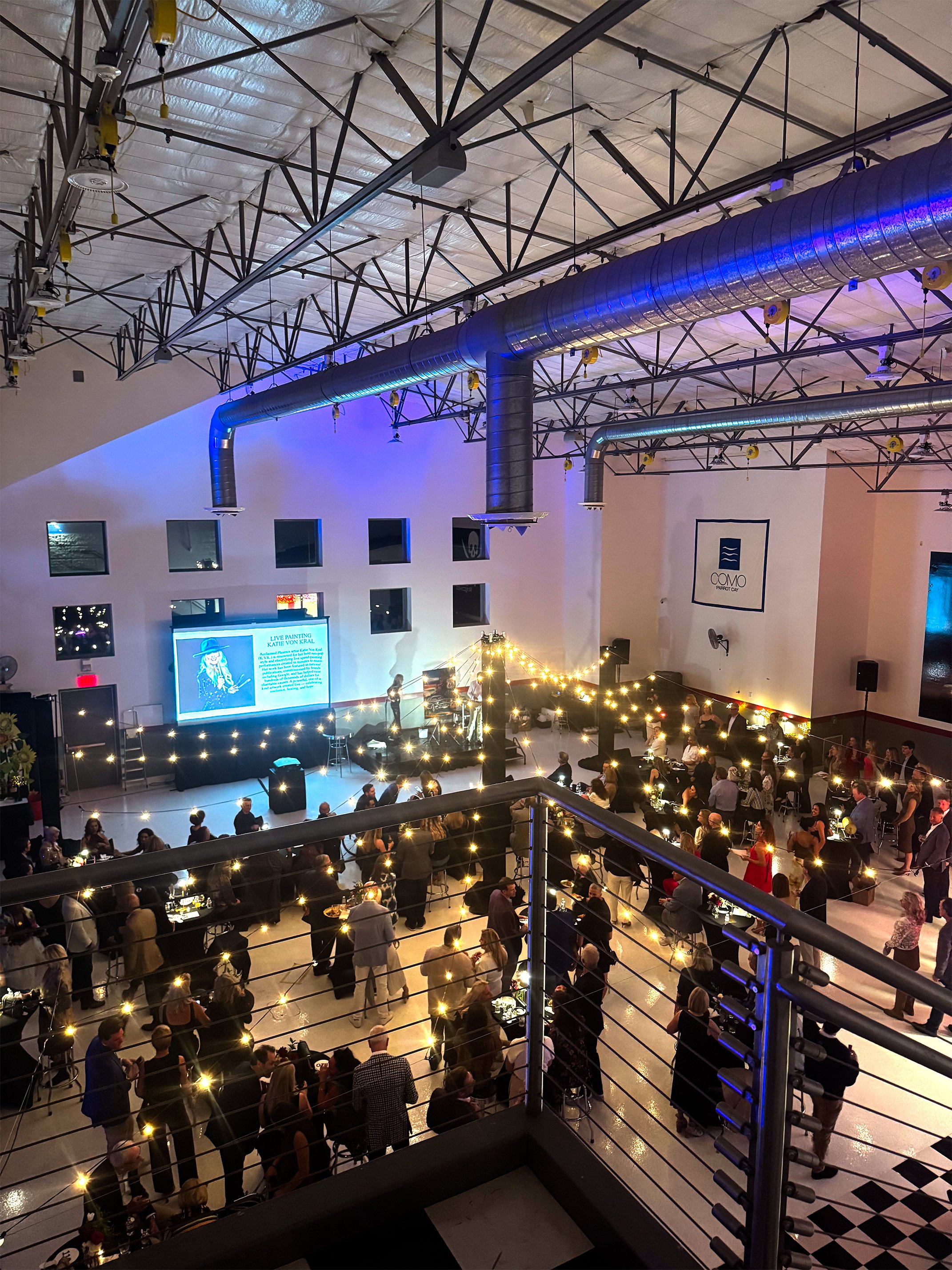 Indoor event with a stage and audience, string lights decorating the space, large air ducts on the ceiling, and a screen displaying a presentation.