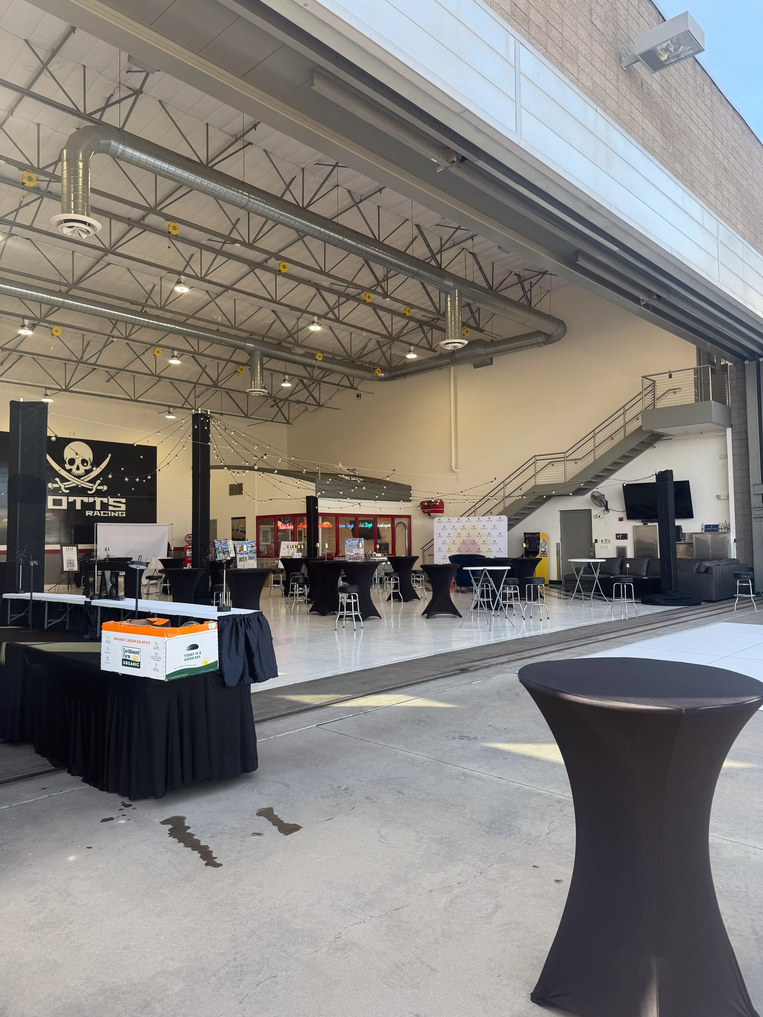 Indoor event space with black high-top tables, some with black tablecloths, and a stage with a banner reading 'OTTS Racing' with a skull logo. There are string lights hanging across the space, and a staircase leading to an upper level. Outside, there
