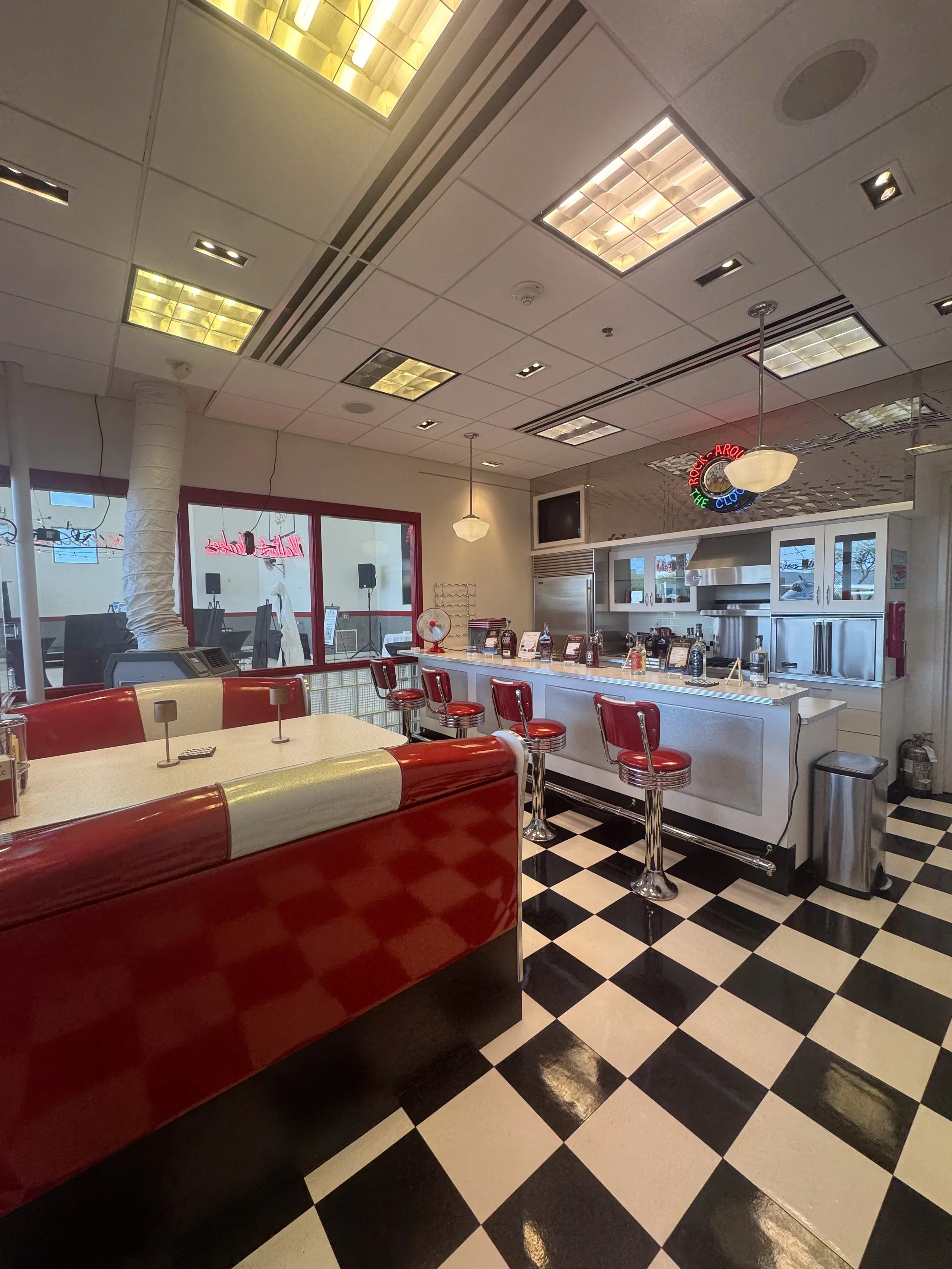 Interior of a retro-style diner with red and white booths, checkered black and white flooring, and a counter with red stools. Neon signs and appliances are visible, with a window reflecting outside.