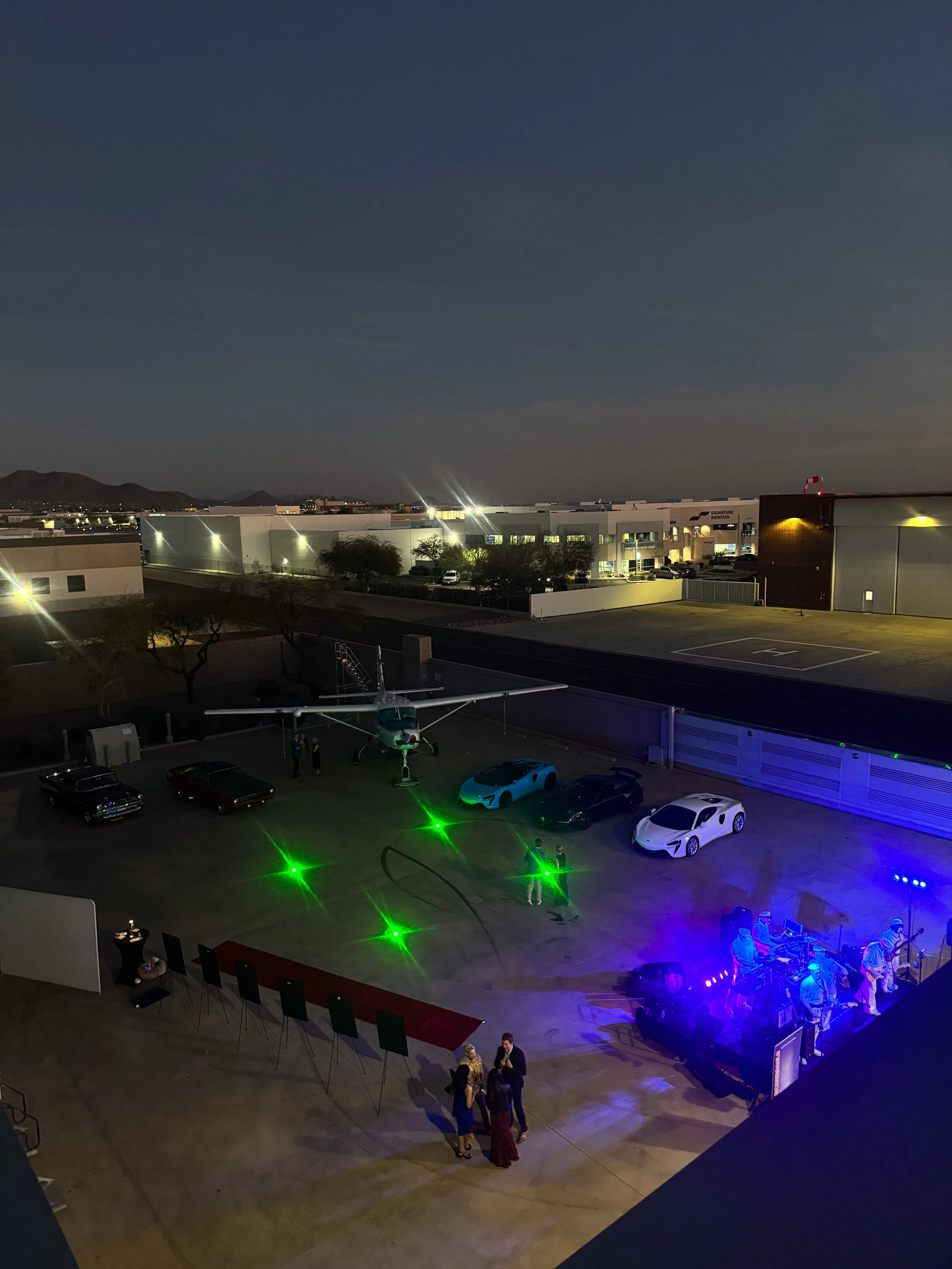 Nighttime scene at a private event with a small aircraft, luxury cars, a stage with a live band, and a few people socializing outdoors.