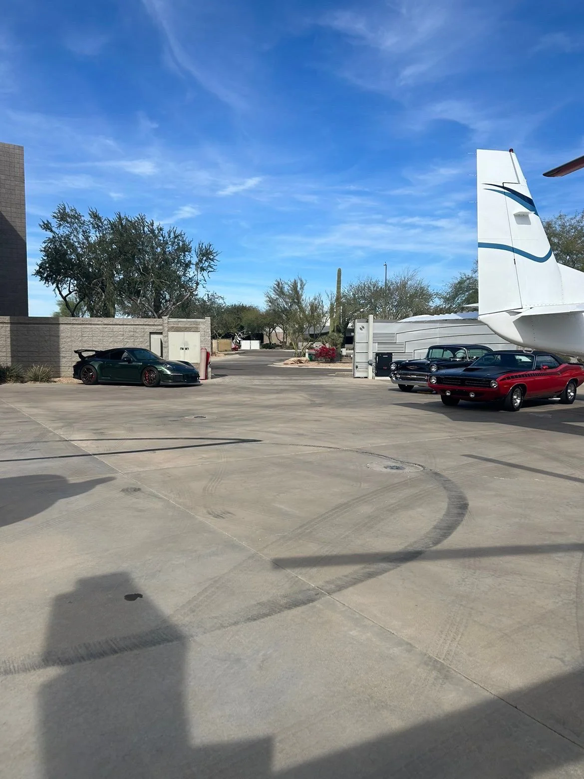 Parking lot with a black sports car, vintage cars, a private jet, trees, and a blue sky.