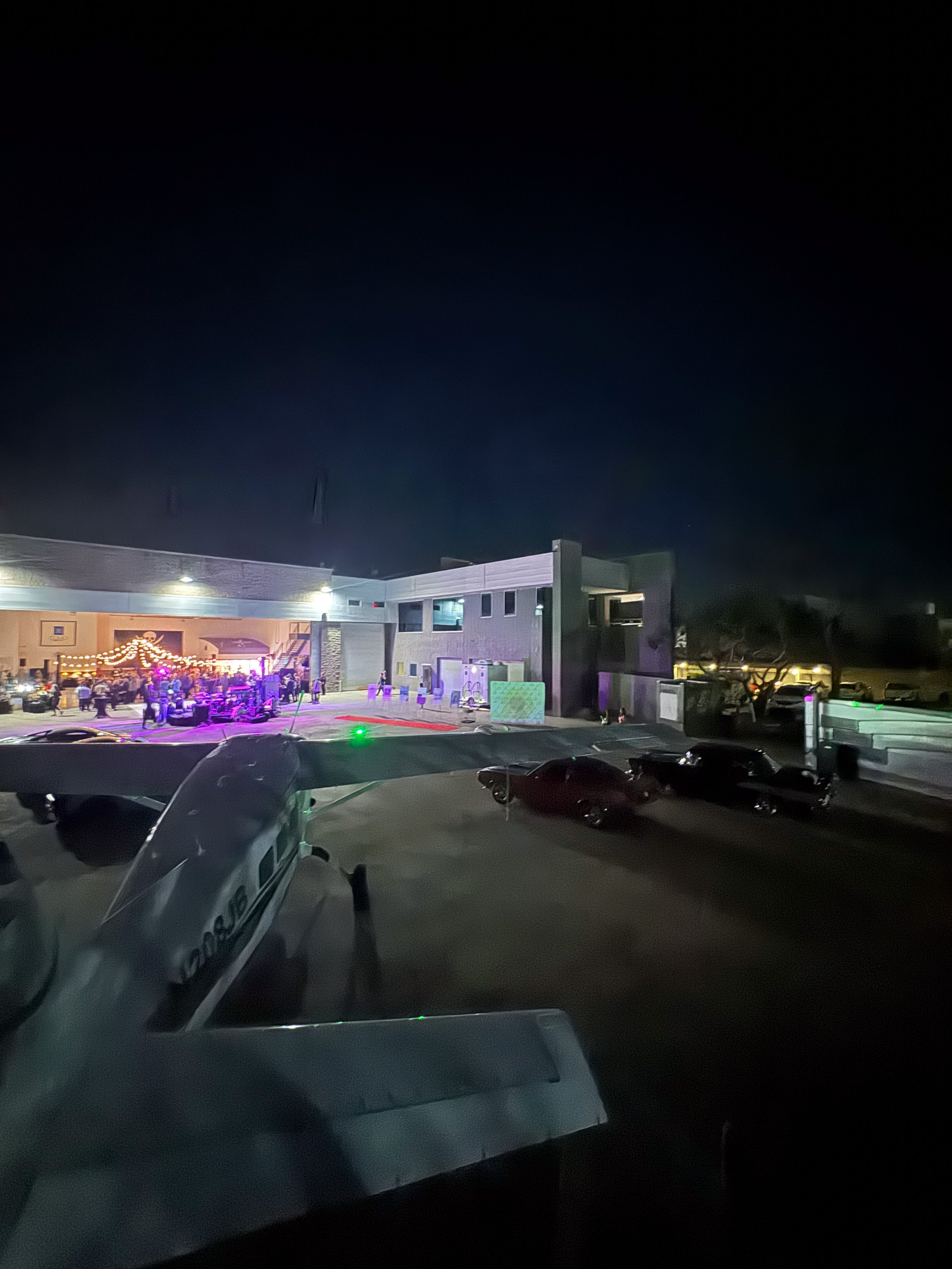 Nighttime scene of a parking lot with a small airplane in the foreground and a building with lights and people in the background.