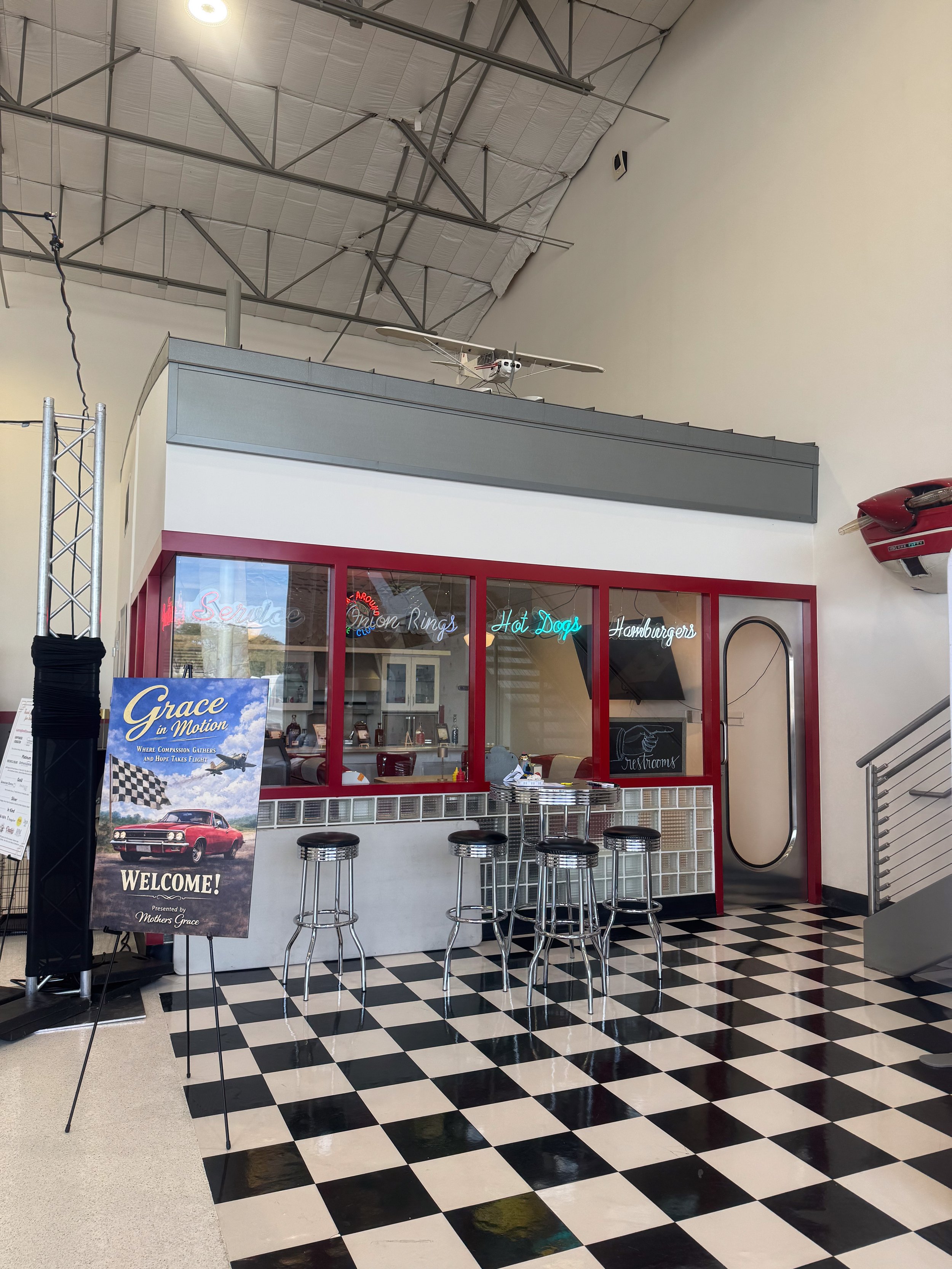 Interior of a vintage-themed diner with a black and white checkered floor, red framed windows, bar stools, neon signs, and a sign welcoming visitors to Grace in Motion.