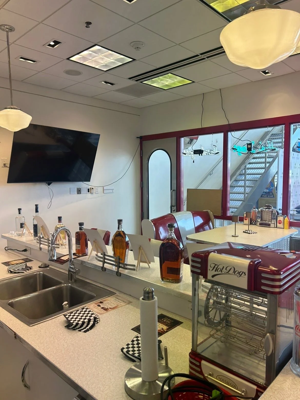 Diner counter with bottles of syrup, hot dog machine, and checkered napkins, with red seating and television on the wall.