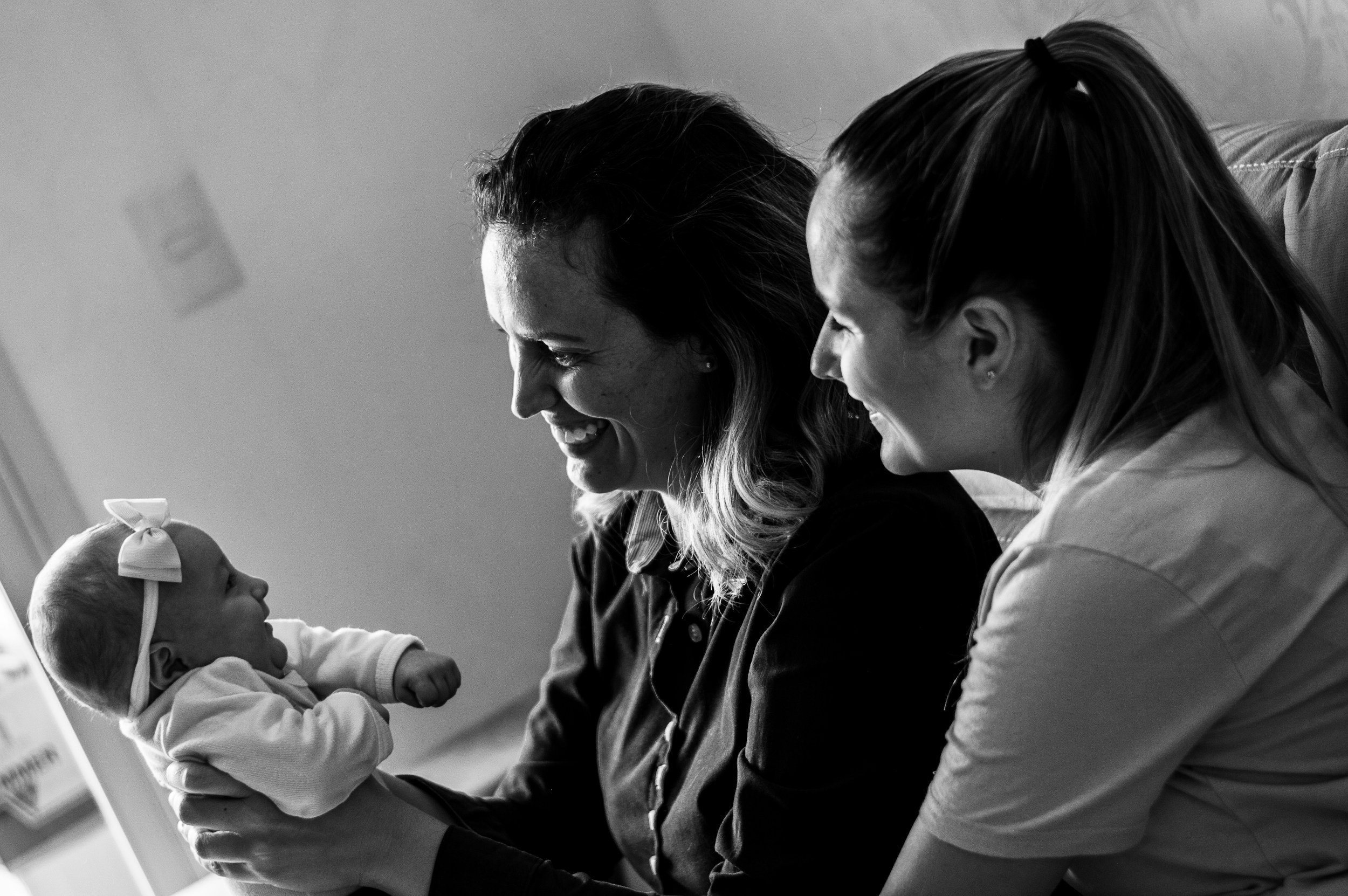A woman, a woman, and a newborn, all smiling and leaning in close, looking at each other.