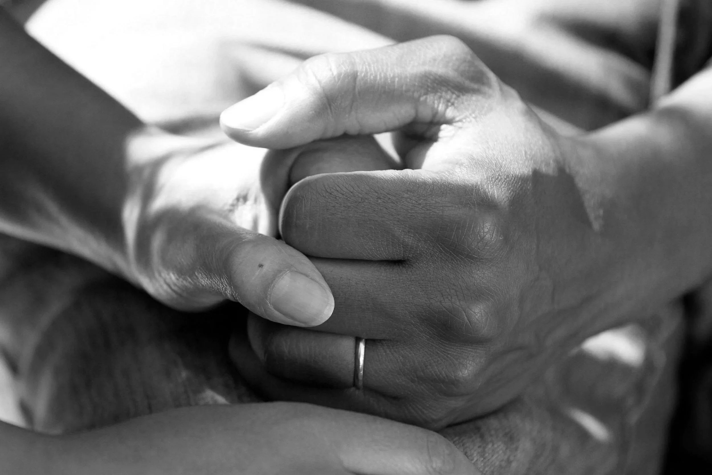 Close-up black and white photo of two hands, one resting on top of the other, with the top hand's fingers gently curled around the fingers of the bottom hand.