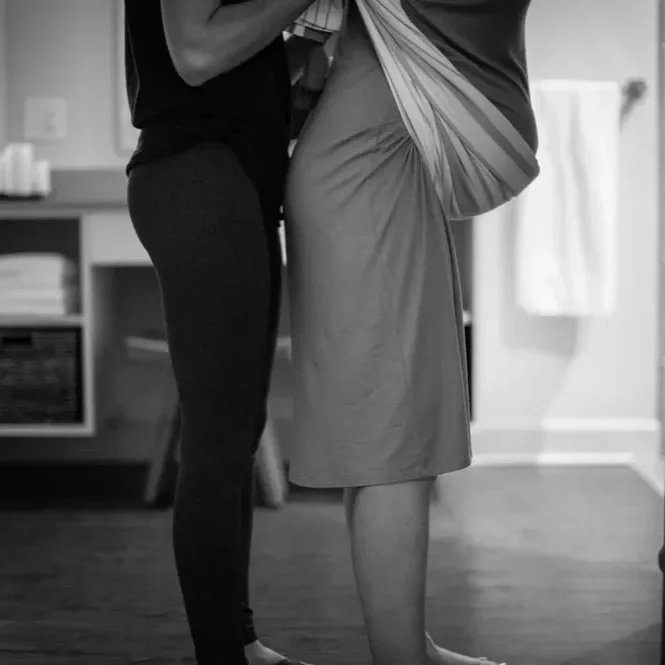 Two people standing close together indoors, with only their lower bodies visible, one in dark pants and the other in a light-colored skirt.