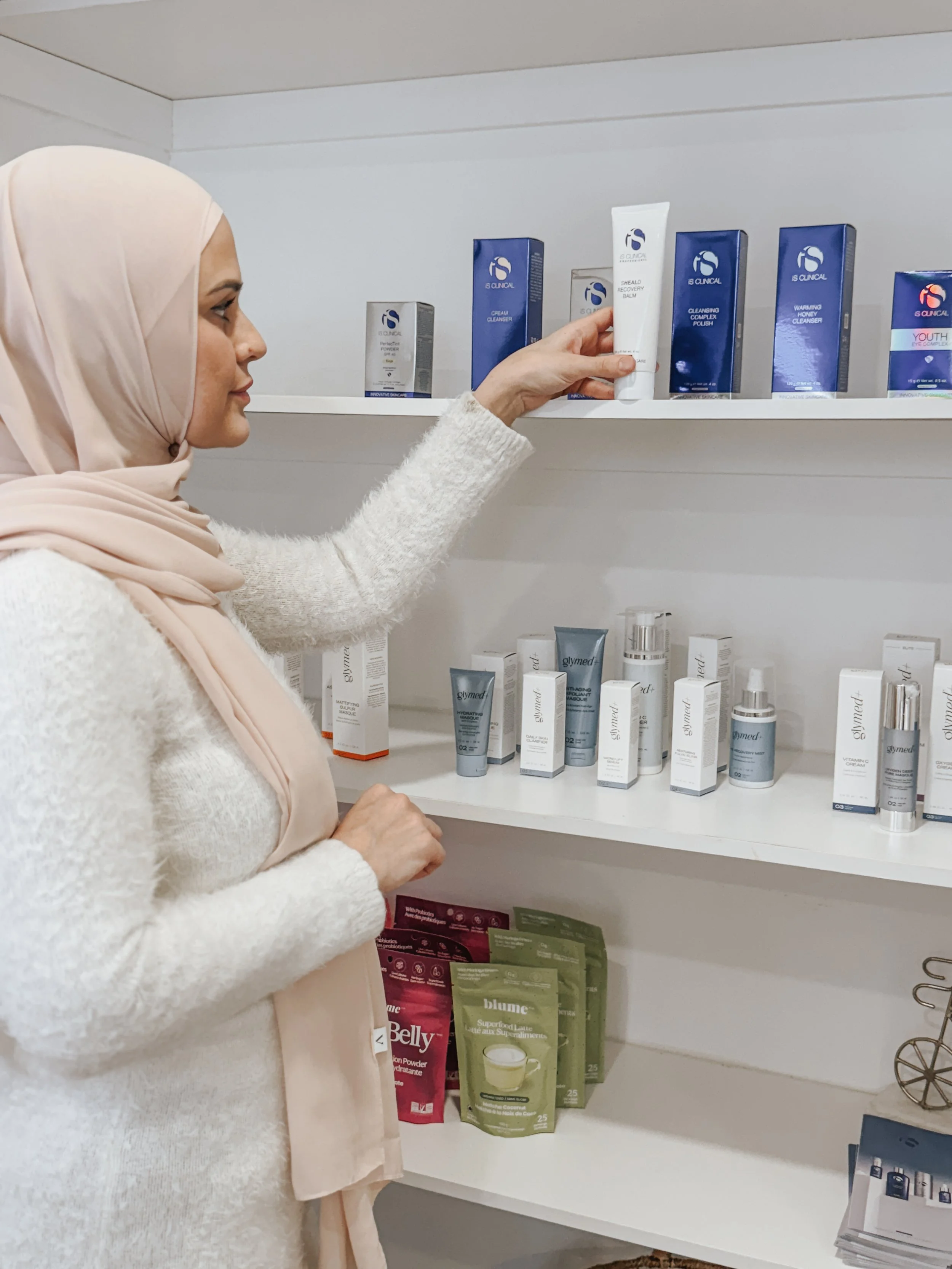 A woman wearing a beige headscarf and a white sweater is browsing skincare products on a white shelf, holding a tube of cream among boxes of other skincare products.