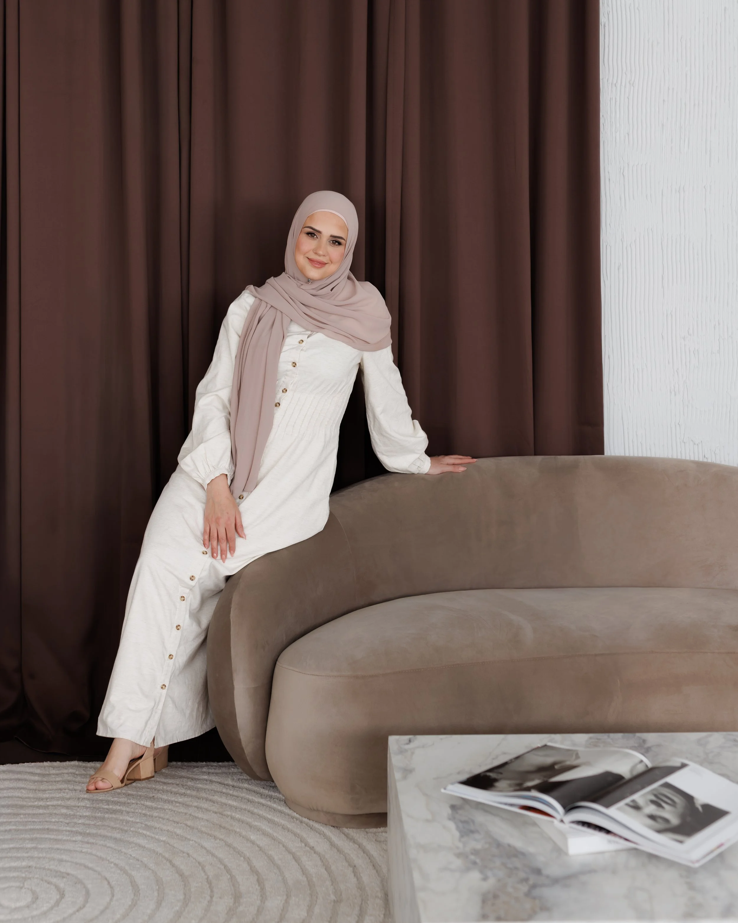 A woman wearing a beige hijab and white button-up dress standing beside a round beige sofa with an open magazine on its marble surface.