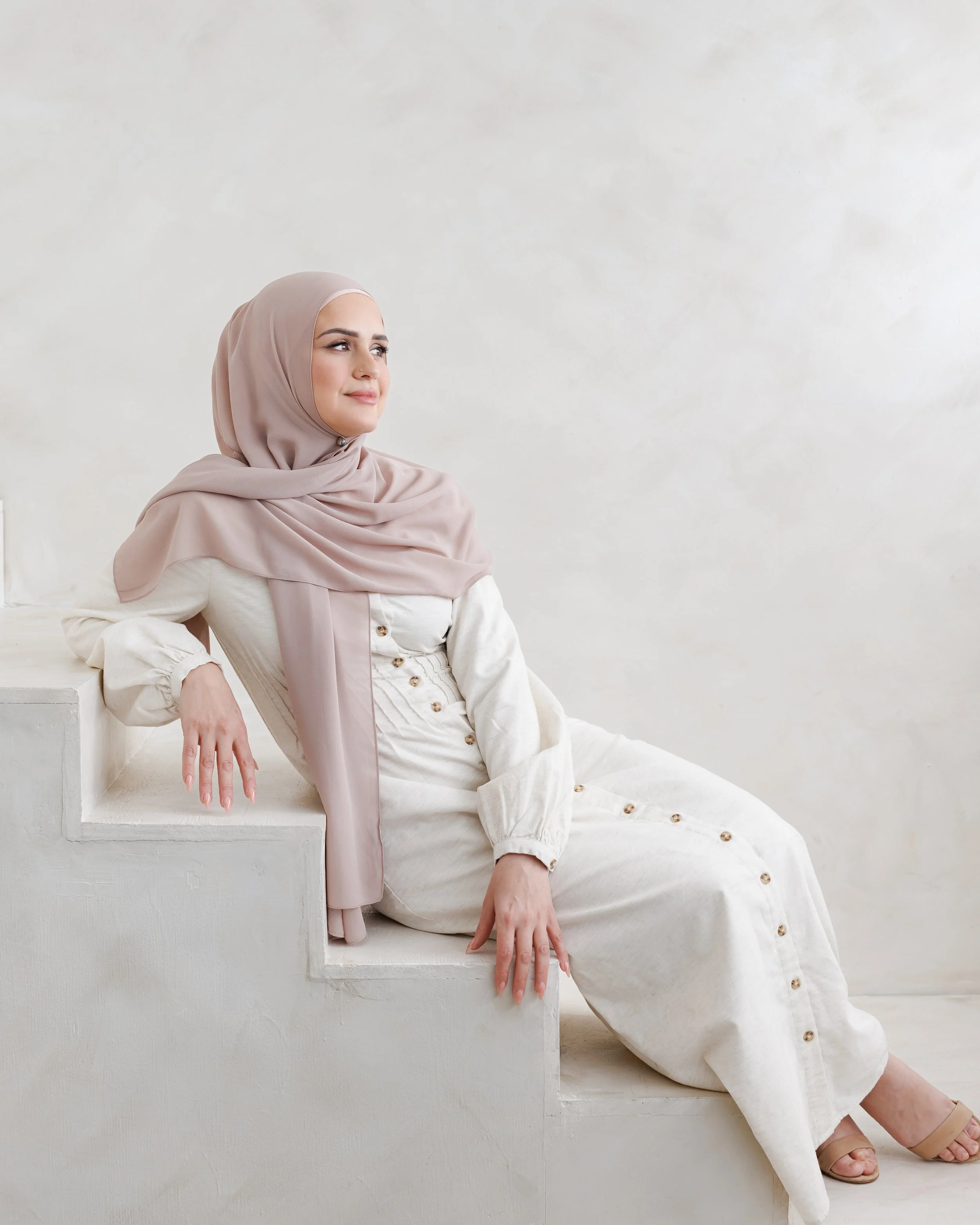 A woman in a light pink hijab and cream-colored outfit sits on a step, leaning back with her arm resting on another step, against an off-white background.