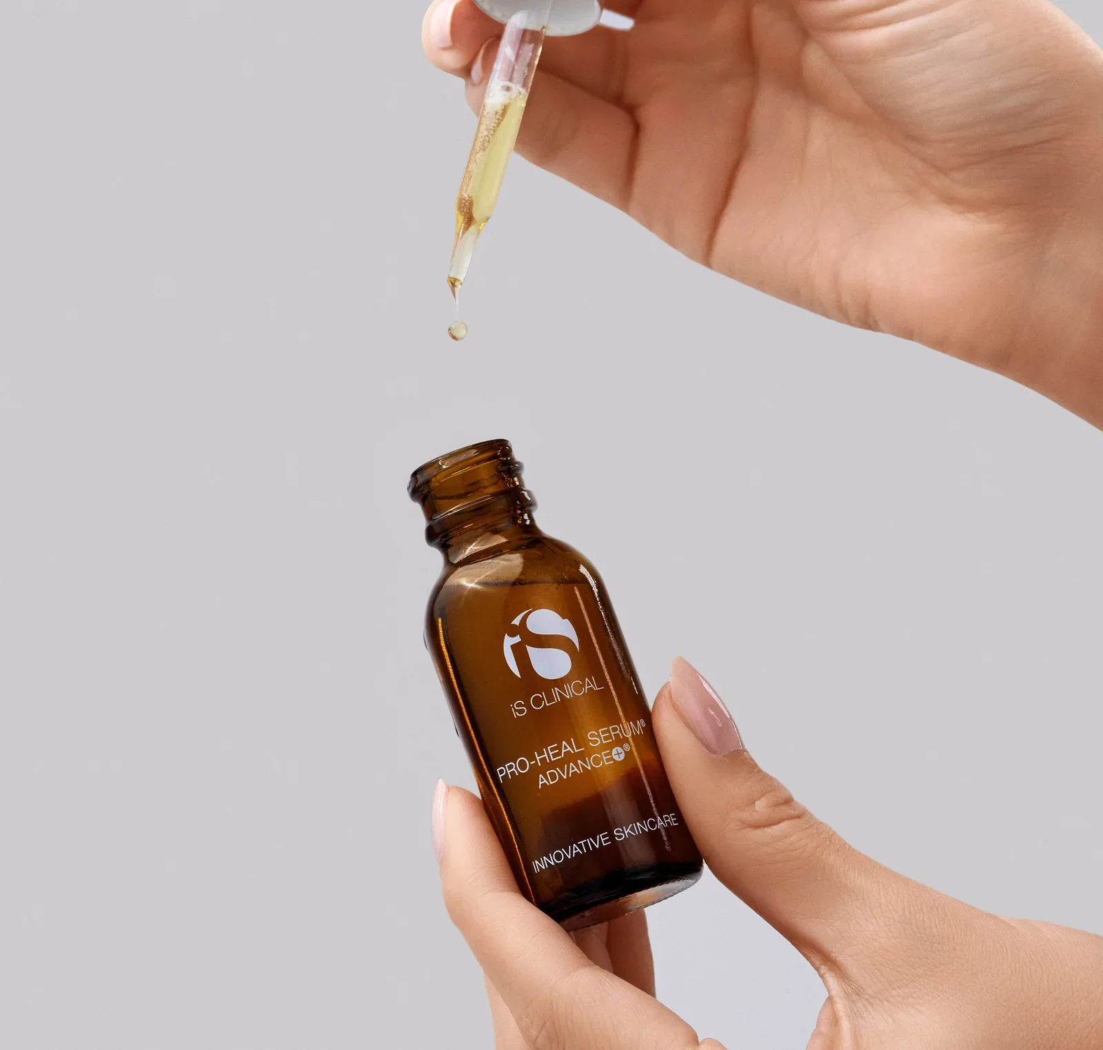 A hand holding a brown glass bottle of skincare serum with a dropper applicator releasing a drop of serum onto a grey background.
