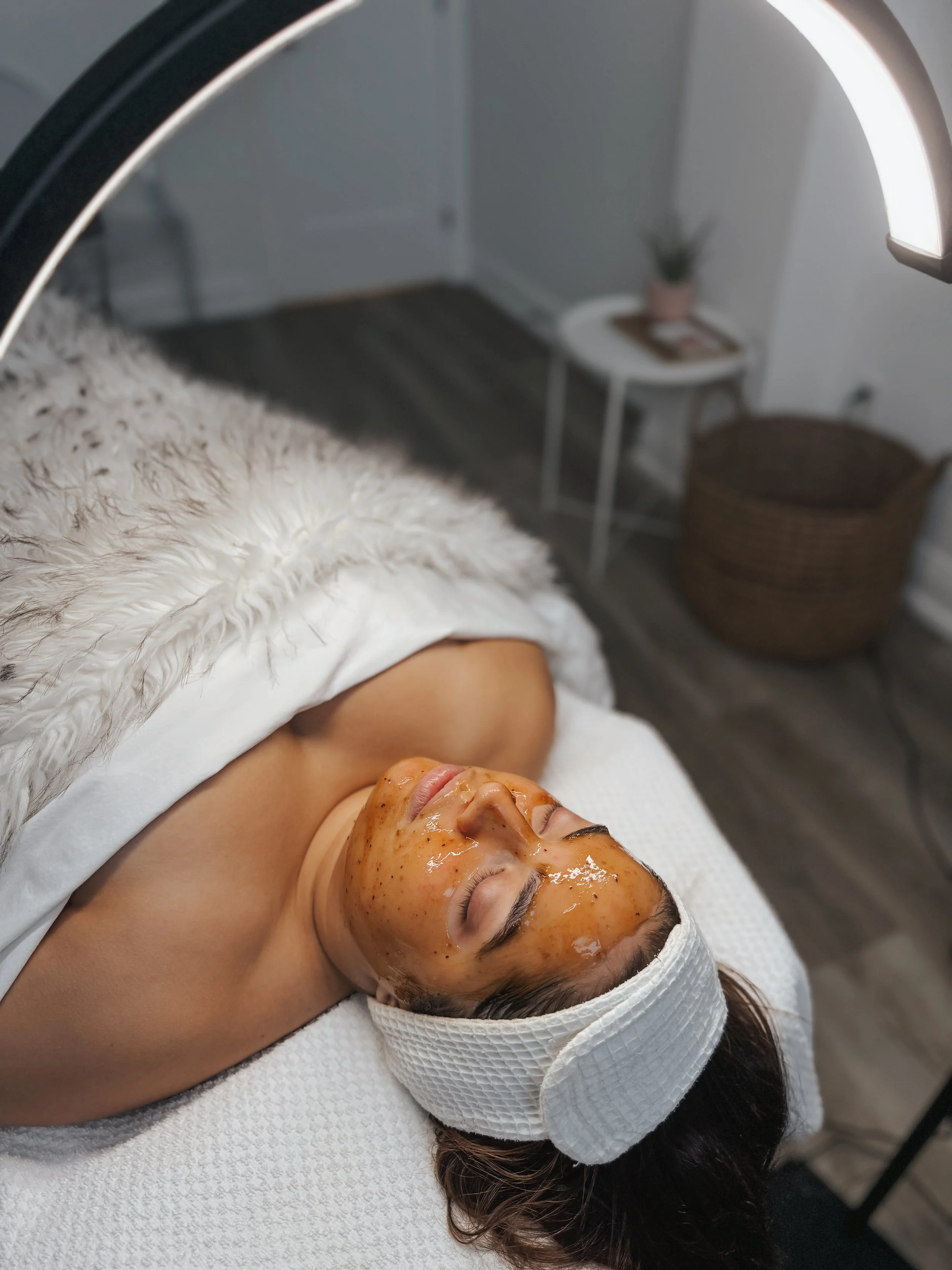 A woman lying on a spa table with a facial mask, wearing a white headband, with her eyes closed and a white towel around her shoulders.