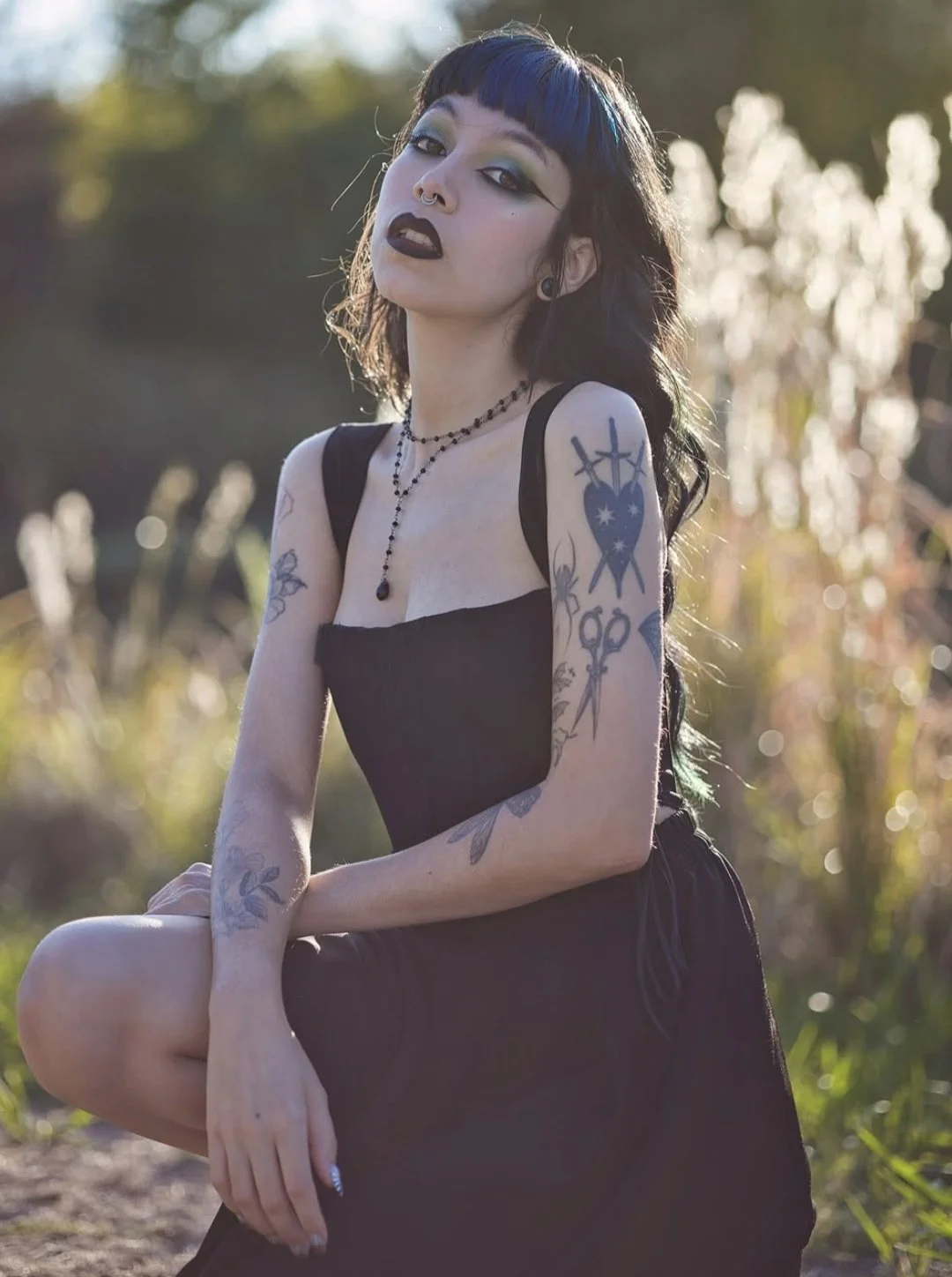 A woman with dark hair, tattoos on her arms, black lipstick, and colorful eye makeup, wearing a black dress and jewelry, posing outdoors with a blurred nature background.