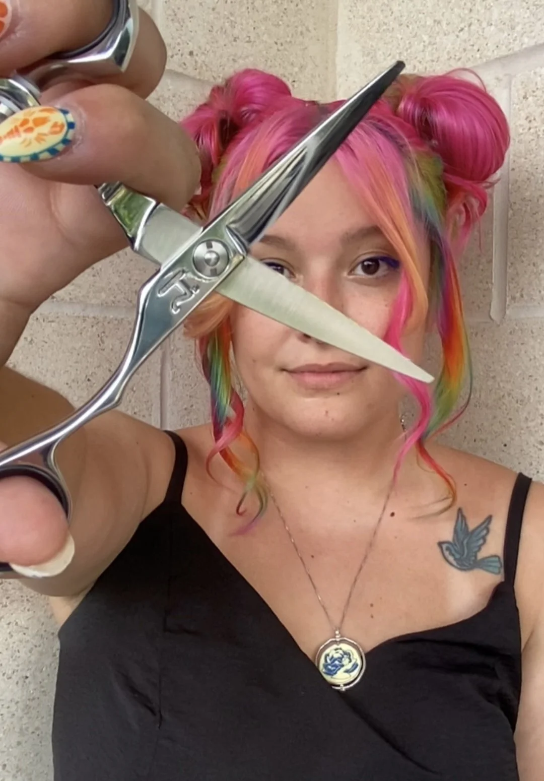 Woman with colorful pink, orange, green, and purple hair styled in buns, holding scissors and a comb, dressed in a black top, standing against a beige tiled wall.